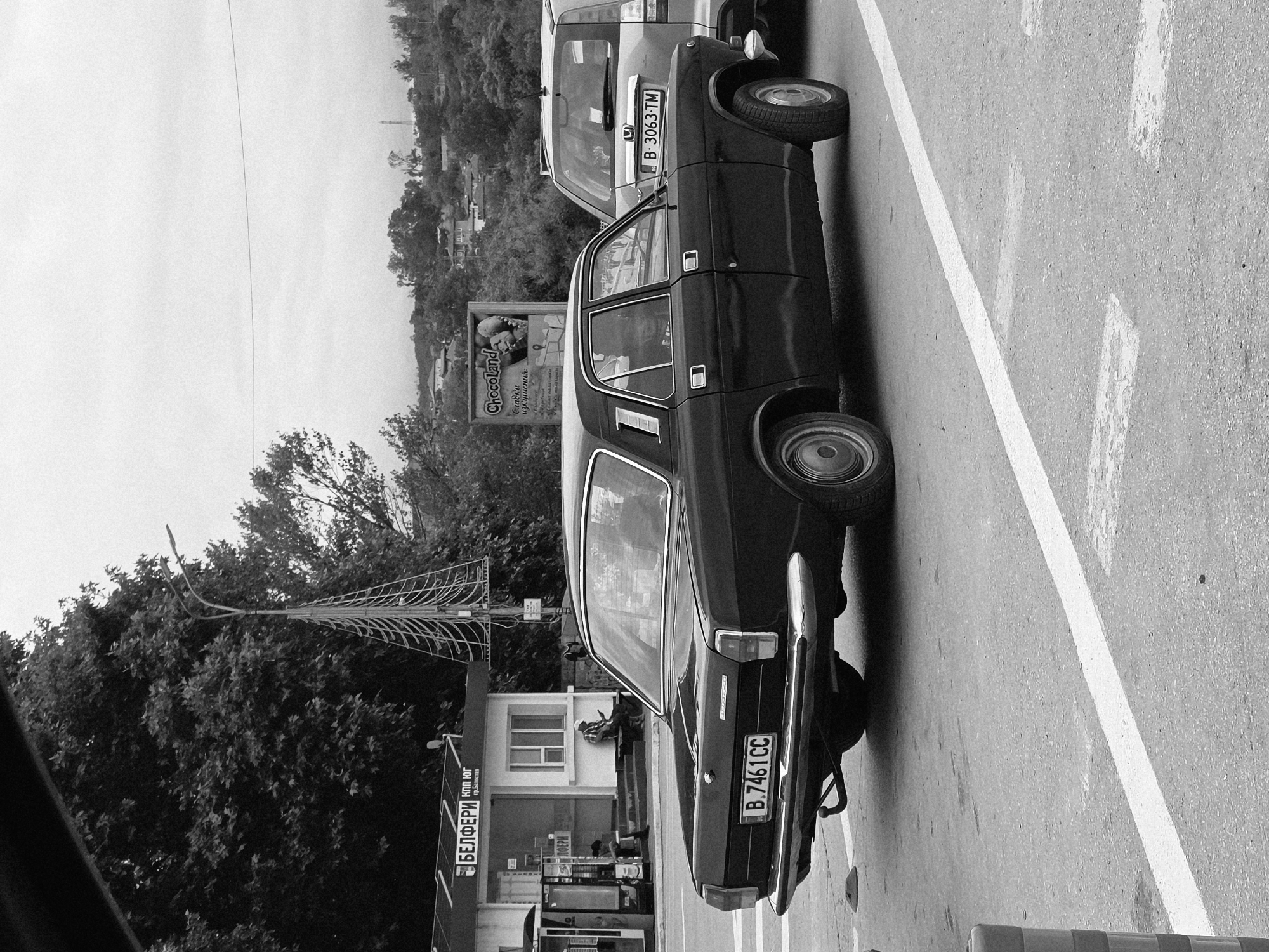 In Beloslav, Bulgaria, a black sedan is parked on an asphalt road, facing away from the viewer. The car's license plate reads "B.7461 CC". To the right of the sedan, a silver SUV is also parked, its license plate visible as "B.3063 TM". Behind the vehicles, a building with signs in Cyrillic is visible, appearing to be a kiosk or small shop. A street light with a unique, geometric design stands near the building. In the background, rolling hills covered in trees and houses suggest a semi-urban or suburban environment under an overcast sky. Two individuals are seated on a bench outside the building. The scene appears to be a street scene during daylight.
