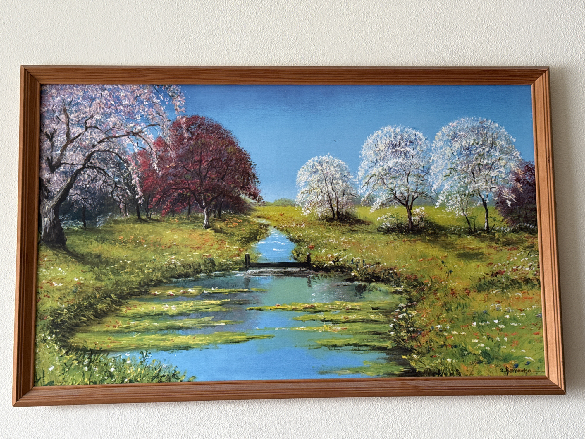 The image is a painting of a serene natural landscape. A narrow, winding river flows through a meadow dotted with wildflowers. On the left side of the river, a cluster of trees with delicate pink blossoms suggests spring. Further into the background, trees with deep red foliage add a touch of dramatic color. On the right side of the river, several trees are covered in white blossoms, creating a soft, cloud-like effect. The sky is a clear, vibrant blue. A small wooden footbridge crosses the river in the middle ground. The painting is framed by a simple wooden border. The painting is signed in the lower right corner with a stylized script that reads "z. Berecsha". There are no people or animals depicted in the painting, and no specific location in Tachov, Czechia, can be identified from the artwork itself.