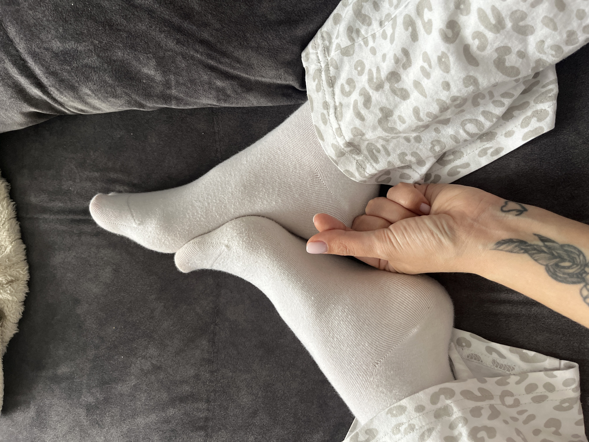 An overhead shot captures a person's lower body, specifically their legs and one arm, in a relaxed indoor setting. The person is wearing light-colored, possibly white or cream, socks that cover their feet and lower legs. They are also clad in pajamas featuring a light gray leopard print on a white background, visible on their upper legs and around the arm.
The left leg is bent, with the foot resting on what appears to be a dark gray, textured blanket or upholstery, possibly a couch or bed. The right leg is also bent, and the person's right hand is gently touching the top of their right foot. The hand has neatly manicured nails with a light, natural-looking polish. A black tattoo, resembling a stylized knot or a heart, is visible on the inner wrist of the hand.
In the background, to the left, a light-colored, fluffy or fuzzy object is partially visible, suggesting another soft textile like a blanket or cushion. The overall ambiance is one of comfort and relaxation, likely indoors, in Chaikovsky, Russia. There is no visible text in the image.