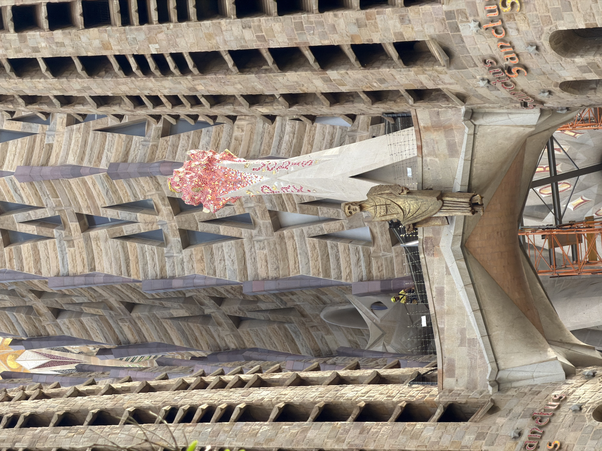 This is a vertical, low-angle shot of the Sagrada Familia in Barcelona, Spain. The iconic basilica's facade is predominantly stone with intricate architectural details. The upper portion of the image shows rows of rectangular openings, creating a textured effect. Below this, the stone construction transitions into a more organic, wave-like pattern with triangular voids.
A prominent feature in the mid-ground is a tall, pointed structure adorned with colorful mosaic tiles in shades of pink, red, and white, interspersed with gold accents. To the right of this, a golden, sculpted figure appears to be carved into the stone. Further to the right, orange construction scaffolding is visible, suggesting ongoing work or maintenance on the building. In the distance, glimpses of the basilica's interior can be seen, with its characteristic organic, tree-like columns and stained-glass windows.
Visible text spelling out "Sanctus" in ornate, orange lettering is present on the stone surface in the lower right of the image, along with small, star-shaped accents. Two figures in yellow safety vests are visible in the mid-ground, seemingly working on the structure or navigating the scaffolding. The lighting suggests it is daytime, with natural light illuminating the stone facade. The overall impression is one of awe-inspiring architecture and a blend of artistry and ongoing construction.