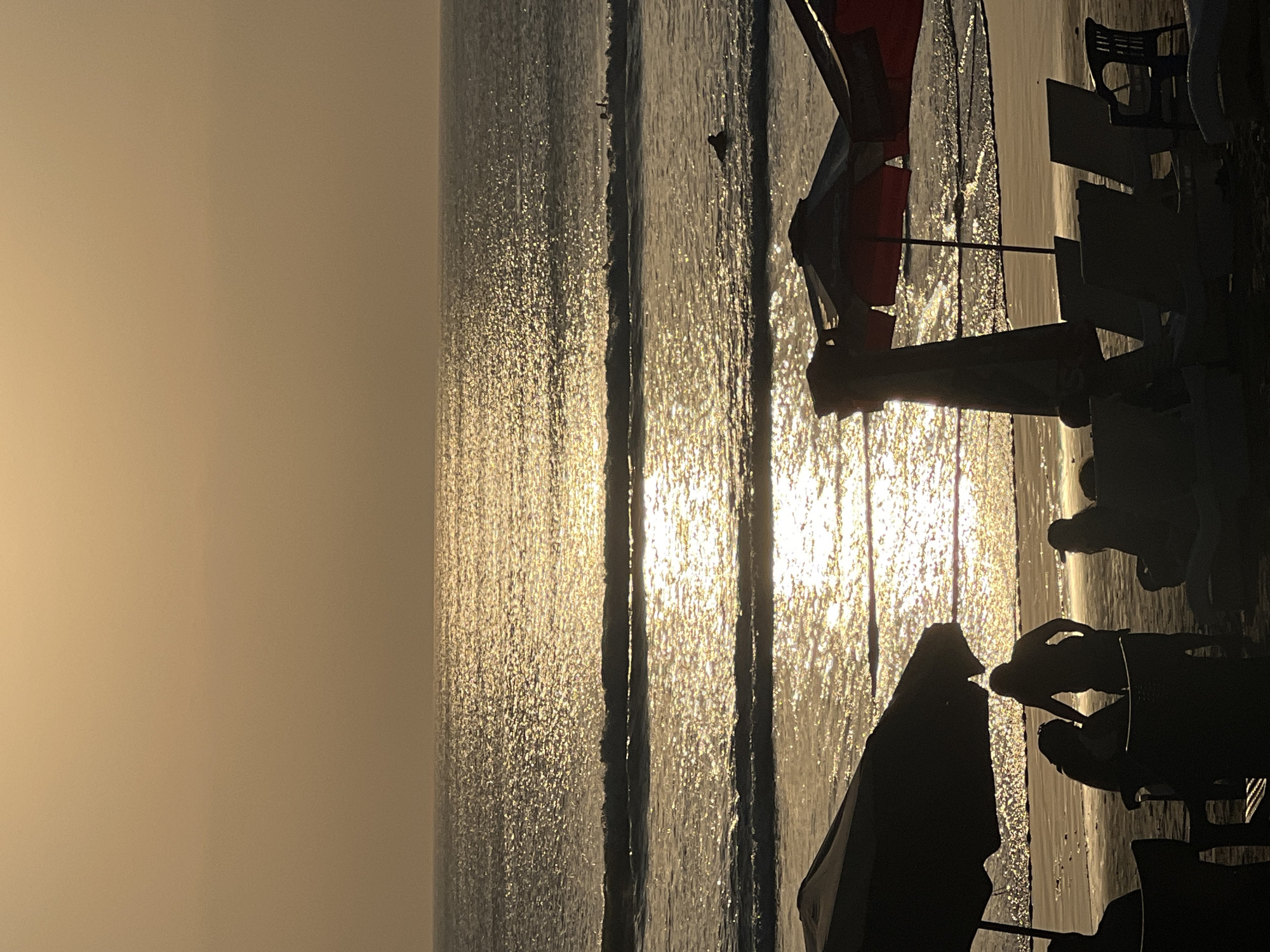 The image depicts a beach scene at sunset in Kuta, Indonesia. The sky is a hazy golden color, and the sun's reflection glitters on the ocean's surface, creating streaks of light across the water. Gentle waves roll onto the shore. In the foreground, silhouetted figures are visible. Two umbrellas, one large and dark, and another partially visible with red accents, are set up on the sand. Several lounge chairs, also in silhouette, are arranged on the beach. In the middle ground of the ocean, a few distant figures can be seen in the water, possibly surfers. The overall impression is a serene, end-of-day atmosphere at a popular tourist beach.