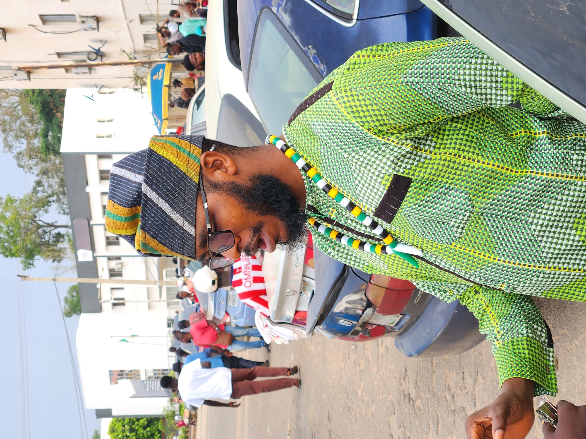 A man with a beard and sunglasses, wearing a green and yellow patterned shirt and a striped hat, stands near a blue car on a paved road in an urban area of Unknown, Unknown. He is wearing a beaded necklace of black, white, green, and yellow beads. In the background, a crowd of people is gathered, some standing near an umbrella with yellow and blue branding. Buildings and trees are visible in the distance.