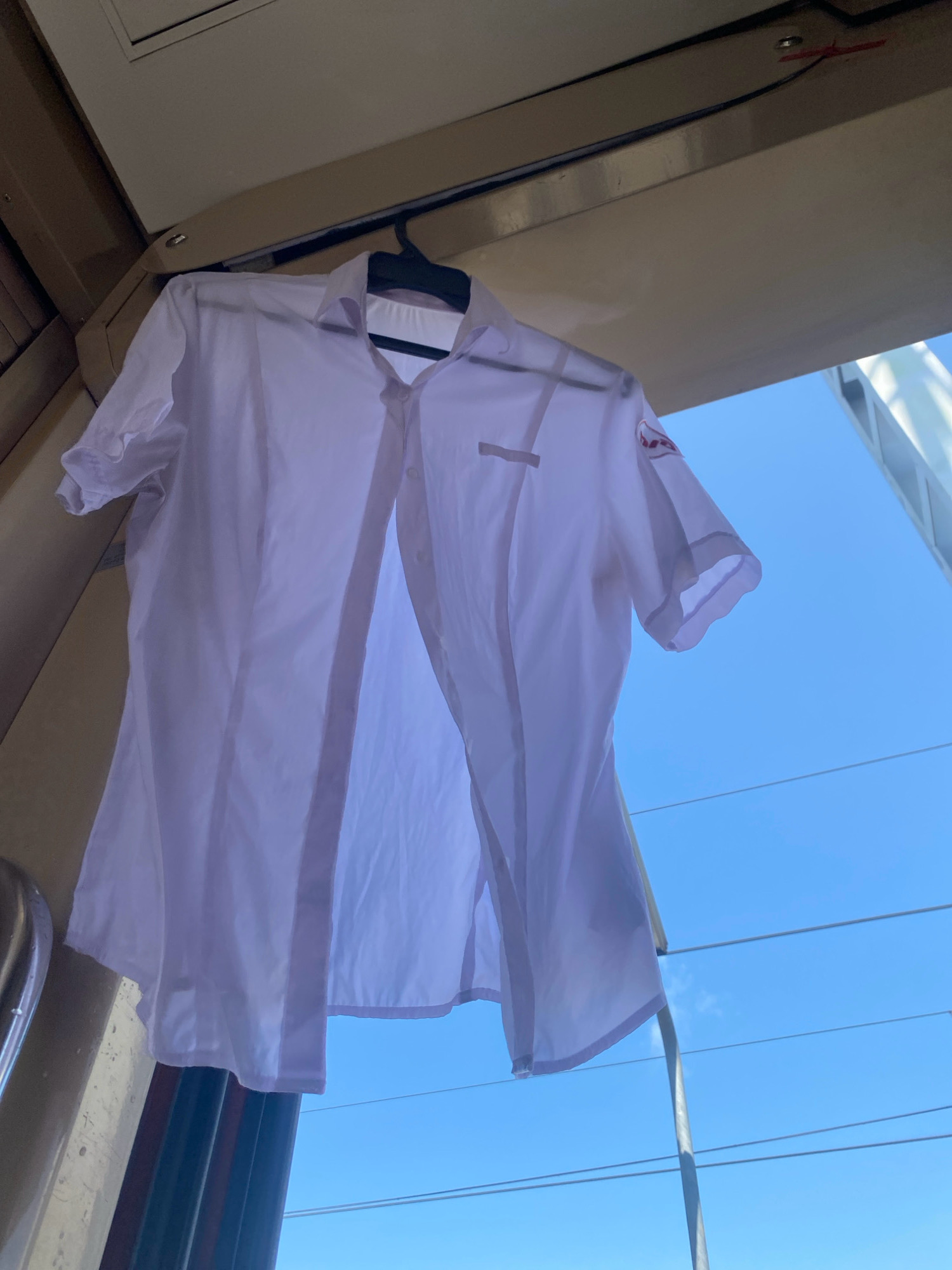A white, short-sleeved shirt is hanging on a black hanger, suspended outdoors. The shirt is open down the front, revealing the inside. The background is a clear blue sky with some power lines visible. The shirt has a small emblem on the left breast. The scene appears to be captured from below, looking upwards.