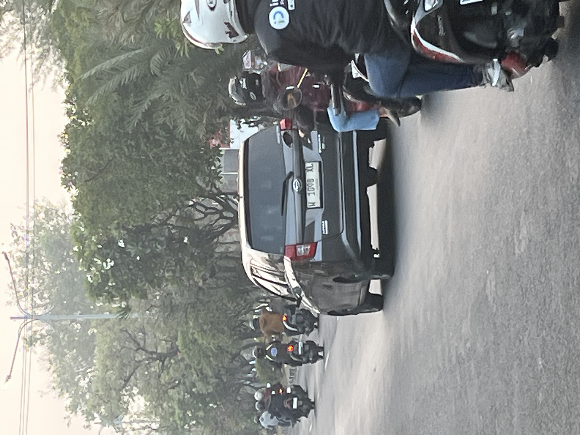The image depicts a street scene in Kludan, Indonesia, characterized by a traffic jam. A dark-colored car, identified by its license plate "W 1098 XL," is prominently positioned in the foreground, obscuring a significant portion of the road. Behind the car, a convoy of motorcycles, each carrying one or two occupants wearing helmets, are stalled. The riders are wearing a variety of clothing, including jackets and casual attire. The environment suggests a suburban or urban setting, with lush green trees lining the road and a street lamp visible in the background. The overall atmosphere is one of congestion, with the vehicles and people appearing to be at a standstill. The lighting suggests it is daytime, possibly early morning or late afternoon, with a soft, diffused light casting over the scene.