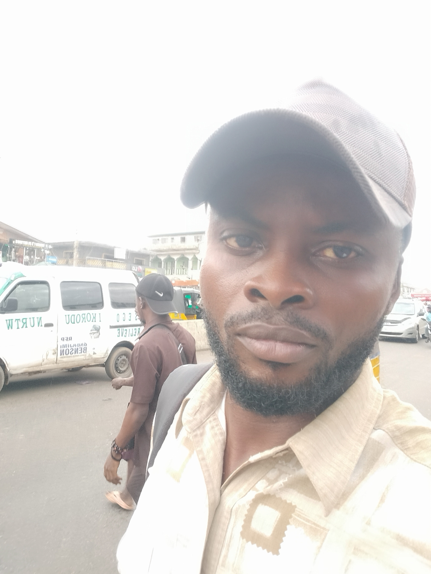 A medium shot photograph shows a Black African male wearing a dark gray baseball cap and a light-colored patterned collared shirt. He has a thick, short beard and is facing the camera. Behind him and to the left, another man wearing a dark baseball cap and a brown shirt walks alongside a white van with green lettering. The lettering on the van says "NURTW IKORODU RED" in English and "BẸ́NSON IMILAGADA" in the Yoruba language. The background features buildings, some with signs, and other vehicles, suggesting an urban street scene. The location is Ikorodu, Nigeria.