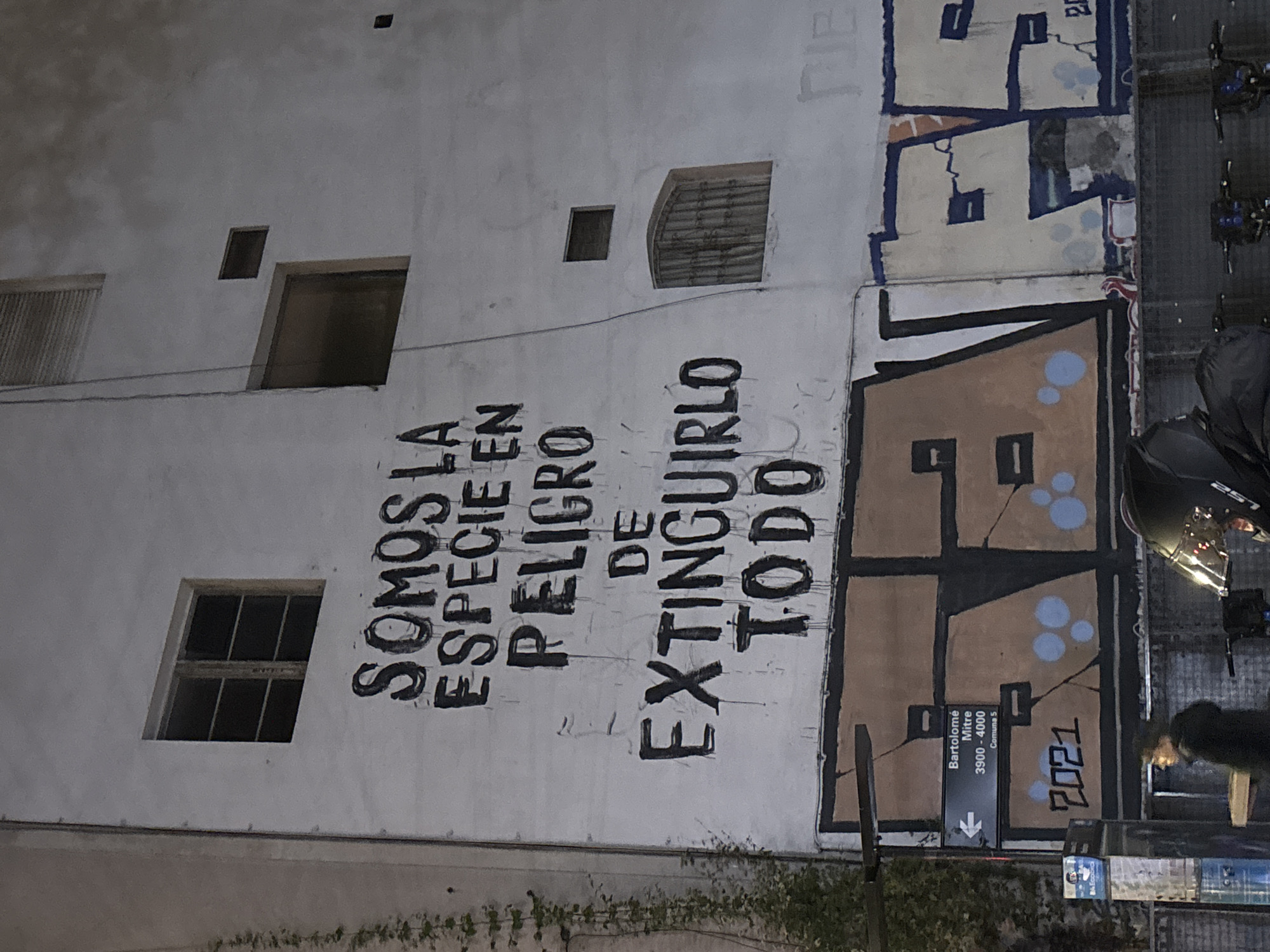 The image depicts the exterior wall of a building in Buenos Aires, Argentina, covered in graffiti and street art. Prominently featured is the Spanish text "SOMOS LA ESPECIE EN PELIGRO DE EXTINGUIRLO TODO," meaning "We are the species in danger of extinguishing everything." Beside this message is a section of abstract graffiti in brown, black, and light blue. A street sign indicating "Bartolomé Mitre 3900-4000 Comuna 5" is visible, locating the scene along Bartolomé Mitre street in the Comuna 5 district of Buenos Aires. The number "2021" is written beneath the street sign, likely indicating the year the graffiti was created. The building wall also features several windows and openings. The presence of bicycles and a partially visible figure suggests an urban environment with pedestrian traffic. The scene is dimly lit, possibly at night.