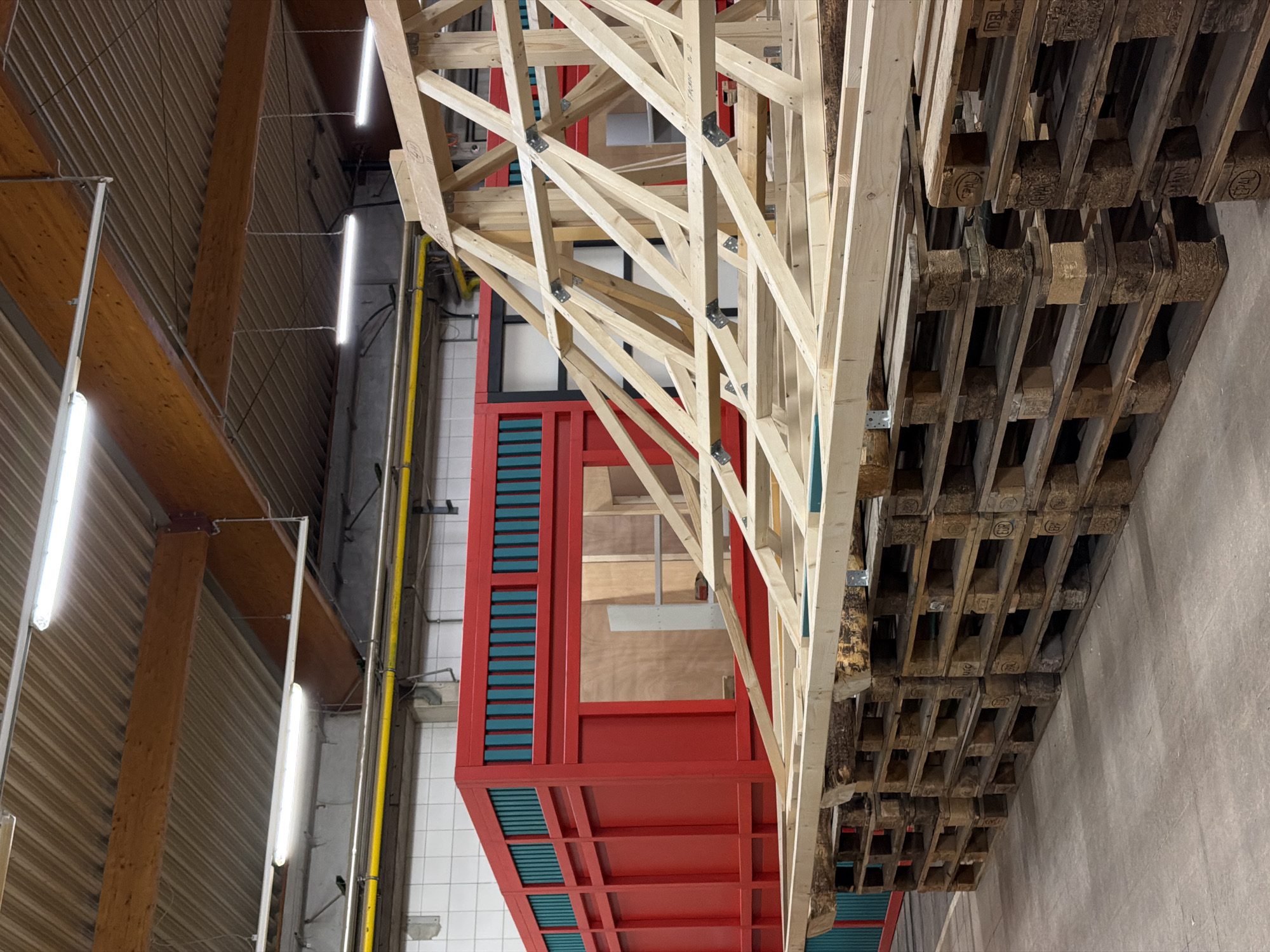 In Leudelange, Luxembourg, a workshop setting features a prominent red and teal structure, possibly a modular building component, situated behind a partially constructed wooden roof truss. The roof truss, made of light-colored lumber, is supported by stacked wooden pallets in the foreground. The environment appears to be an indoor industrial space, evidenced by the concrete floor, exposed ceiling beams, and industrial lighting fixtures. The background includes white tiled walls and a yellow pipe running along the ceiling. No people are visible in the image.