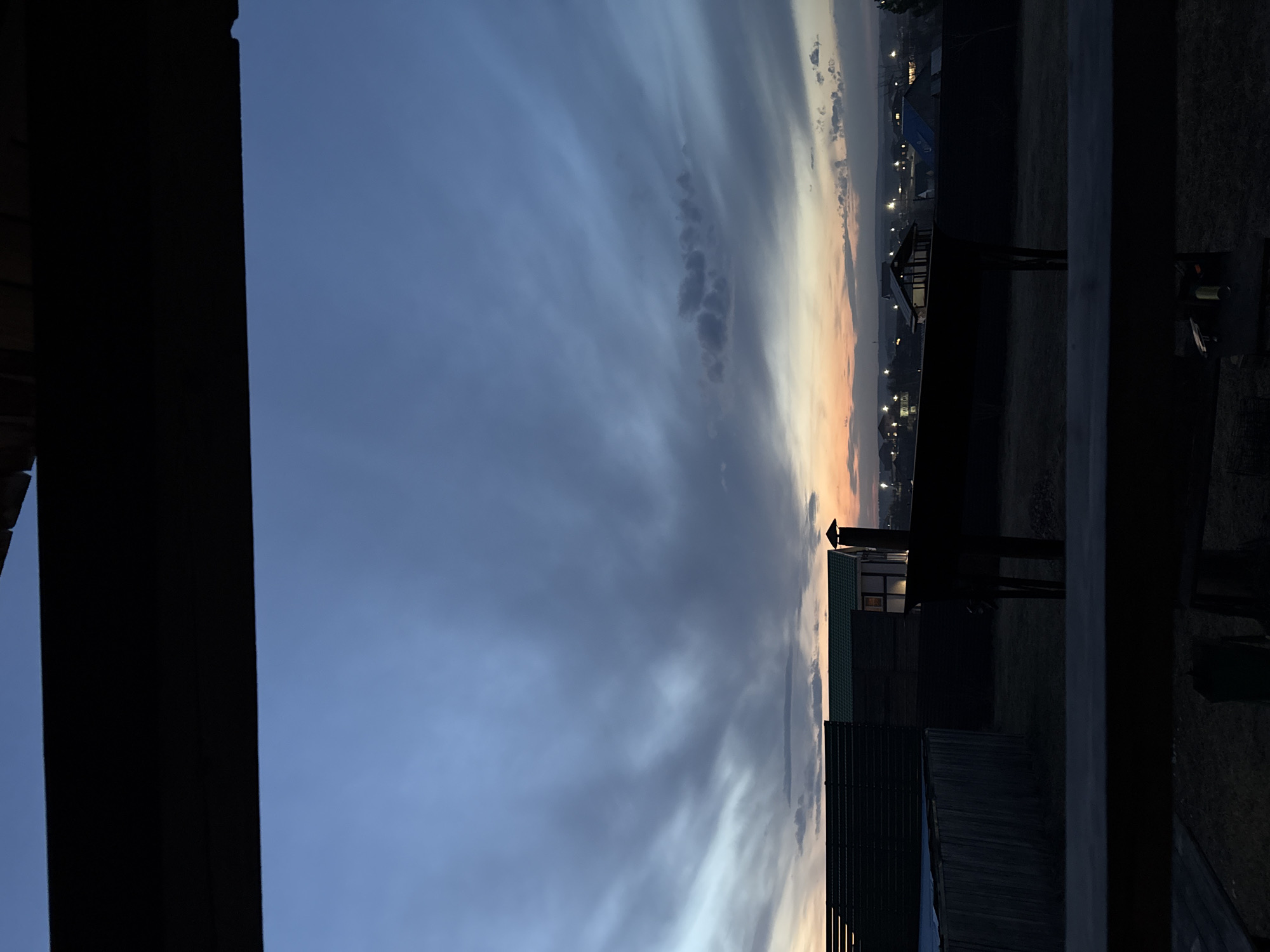 The image captures a twilight or dusk scene from Бугачёво, Russia. The sky is predominantly blue-gray with streaks of orange/yellow near the horizon. Foreground elements include architectural structures or barriers, possibly parts of buildings or fences, that frame the view of the landscape. Distant buildings and street lights are also visible. The image appears to be taken from an elevated position, or perhaps from behind an enclosure.