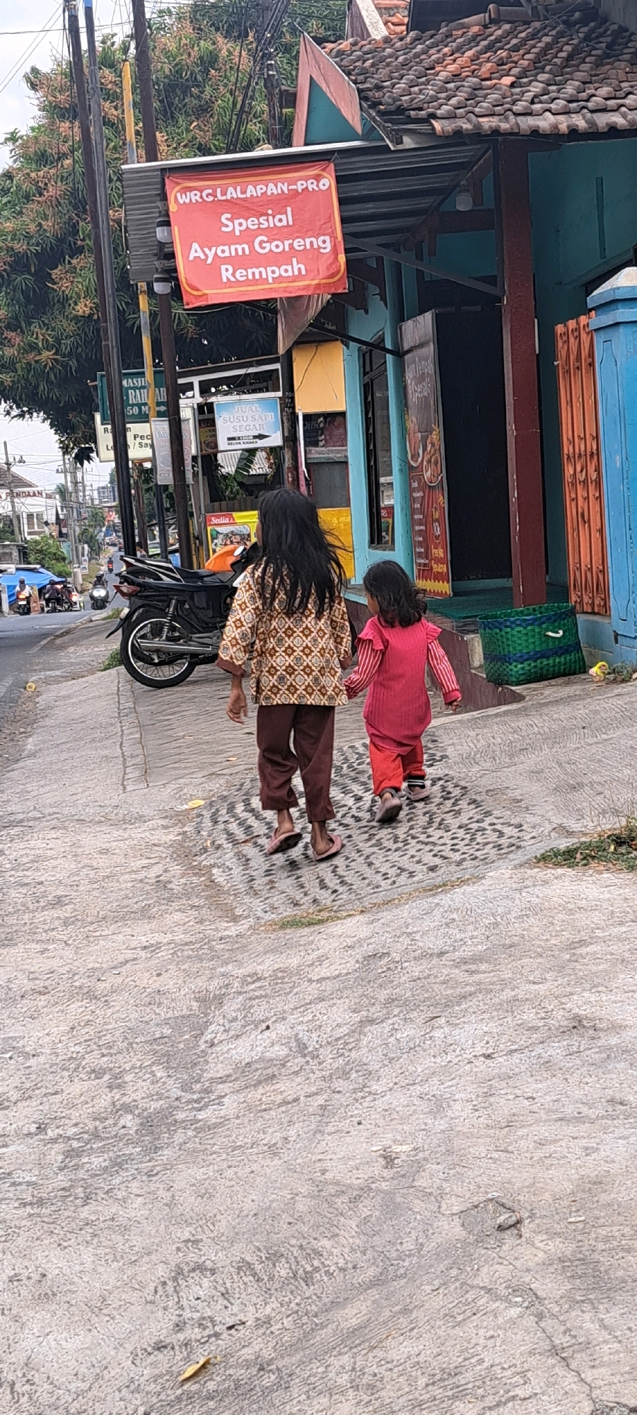 Two young girls, viewed from behind, walk hand-in-hand on a cracked concrete sidewalk. The taller girl on the left wears a patterned long-sleeved shirt and brown pants, with pink flip-flops. The shorter girl on the right wears a pink long-sleeved shirt and matching pants, with dark shoes. They are walking away from the viewer, towards a street where motorcycles and a car are visible.
Behind the girls, a building has a red banner that reads "WRC LALAPAN-PRO Spesial Ayam Goreng Rempah," indicating it is a restaurant specializing in spiced fried chicken. The building features a blue facade and a tiled roof. A green woven basket is placed near the entrance. The environment appears to be a street scene in a town or city, with utility poles and trees lining the sidewalk. The overall lighting suggests it is daytime. The location is identified as Pandaan, Indonesia.