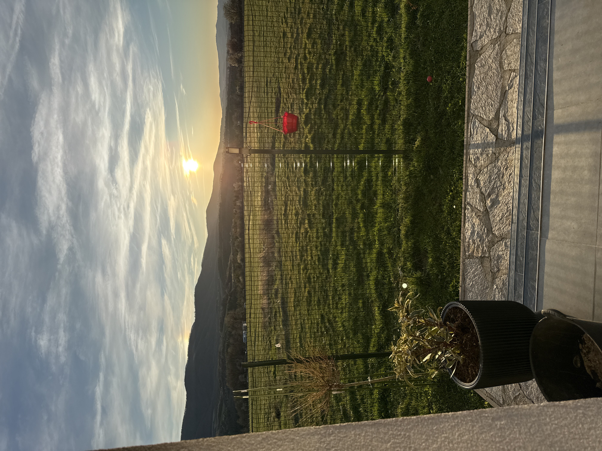 The image shows a view overlooking a green, grassy field with a chain-link fence in the middle ground. A red object, possibly a basket, hangs from the fence. In the background, mountains are silhouetted against a bright sunset sky with wispy clouds. To the right, there is a stone-tiled area and a black plant pot with a plant inside. The image appears to be taken from a balcony or raised platform. I cannot identify the specific location in Sofia, Bulgaria, without additional information.
