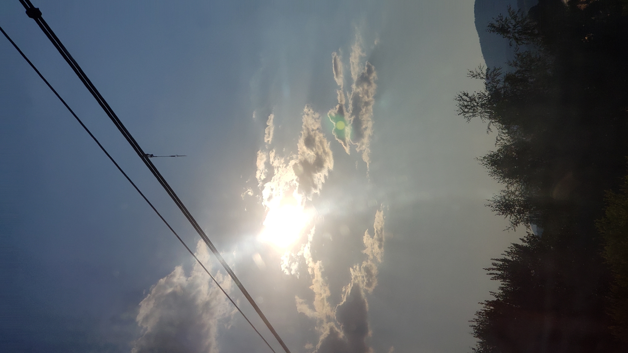 The image depicts a bright sky with scattered clouds and the sun shining through, casting light rays. Two power lines run diagonally across the upper portion of the frame. Below the sky, a silhouetted line of trees forms the lower part of the scene, with a hint of a hill or mountain in the far distance. The overall impression is of a daytime outdoor scene, possibly taken in Liptovský Michal, Slovakia, given the context provided. There are no people or animals visible in the image. The scene is static and there are no discernible actions or interactions taking place. The environment appears to be natural, with vegetation and a hilly landscape.