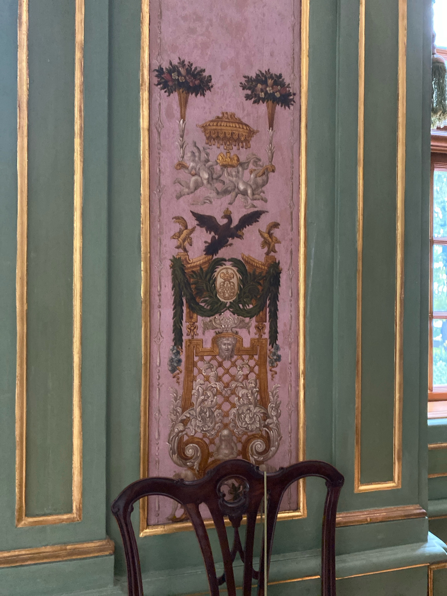 The image captures a detail of the interior of the Stroganov Palace in Saint Petersburg, Russia. The focus is on a wall decorated with elaborate trompe-l'oeil painting. The wall is divided into vertical panels, with the central panel featuring a large, ornate crest. This crest includes an eagle, a crown, cherubs, floral arrangements in cornucopias, and elaborate scrollwork. The background of this central panel is a light pink with a subtle damask pattern. Flanking the central panel are wider, vertical sections of wall painted a muted green, accented by thin gold trim. The lower portion of the image shows the top of a dark wooden chair with a curved back, positioned in front of the wall. A window with visible panes and greenery outside can be seen to the right of the decorated wall. The overall impression is one of classical, opulent interior design.
