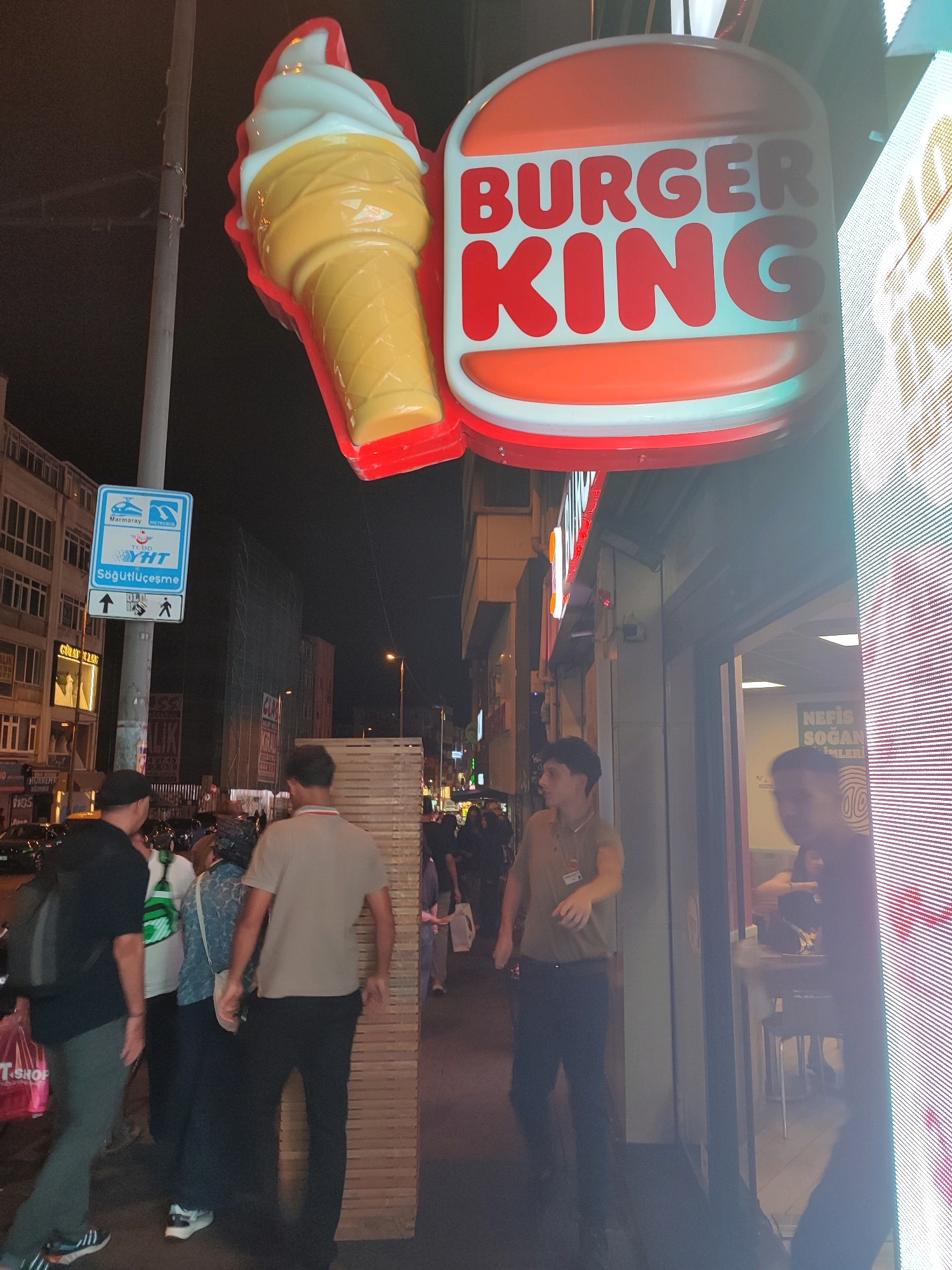 This image depicts a Burger King restaurant located in Kadıköy, Turkey. The signage features the iconic Burger King logo with "BURGER KING" written in red letters against a white background, flanked by an illuminated ice cream cone. The exterior of the establishment is visible, with a glass entrance and a large, brightly lit LED screen on the right side.
Several individuals are present. In the foreground on the left, a person wearing a black t-shirt and dark pants walks past a red shopping bag. To their right, a group of people are walking in the same direction. One individual, wearing a light brown collared shirt and black pants, appears to be an employee and is facing away from the camera, gesturing with their right hand. Further inside the establishment, through the glass, another person is visible seated at a table.
The environment is an urban street at night, characterized by artificial lighting from the restaurant and surrounding buildings, streetlights, and the LED screen. A pole with a sign indicating public transportation routes is also visible. The overall impression is a bustling evening scene at a fast-food restaurant in a city.