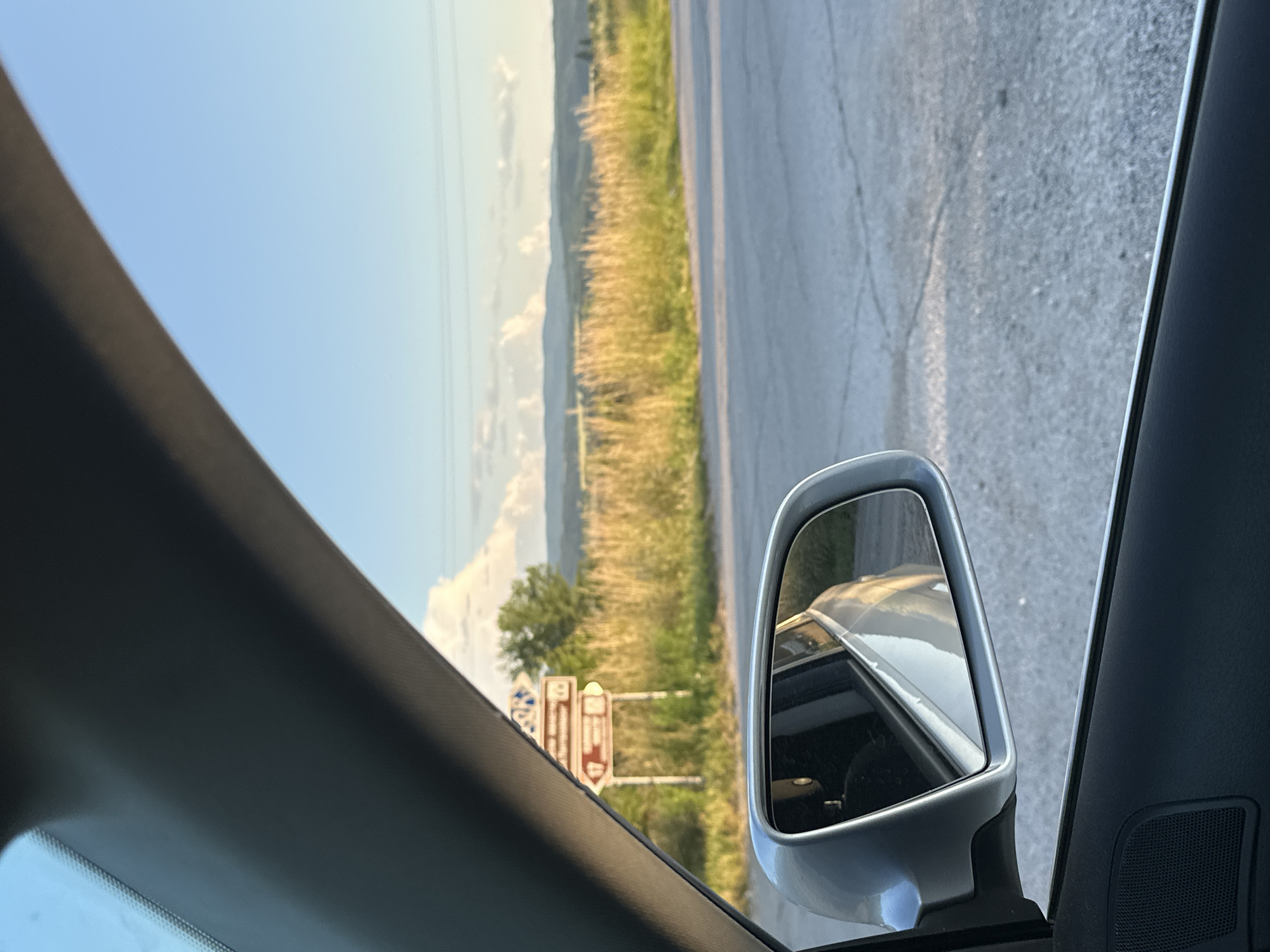 The image depicts a static scene observed from the interior of a vehicle, specifically from the driver's side or front passenger seat looking forward and slightly right. The A-pillar and a portion of the vehicle's interior roof lining are visible in the upper left, while the side mirror and a section of the door panel are prominent in the lower left.
Through the windshield and reflected in the side mirror, a rural landscape unfolds under a clear sky with scattered cumulus clouds, illuminated by the warm, low light of a late afternoon or early evening. The terrain includes a light-colored asphalt road with visible cracks in the foreground. To the right of the road, a wide expanse of dry, golden-yellow grass stretches towards a line of green deciduous trees. Beyond the trees, rolling hills and distant mountains are visible under the horizon.
A brown and white road sign on a post stands blurred in the mid-ground, partially obscured by the vehicle's interior structure. Although details are indistinct due to focus and distance, two directional arrows are visible, one pointing left with the number "17" and another pointing right with the number "88" or similar numeric characters. Overhead, faint power lines traverse the distant sky. The vehicle's exterior, reflected in the side mirror, appears light-colored, possibly silver or white.
The scene is located in Debelets, Bulgaria. No individuals or specific actions are discernible within the frame.