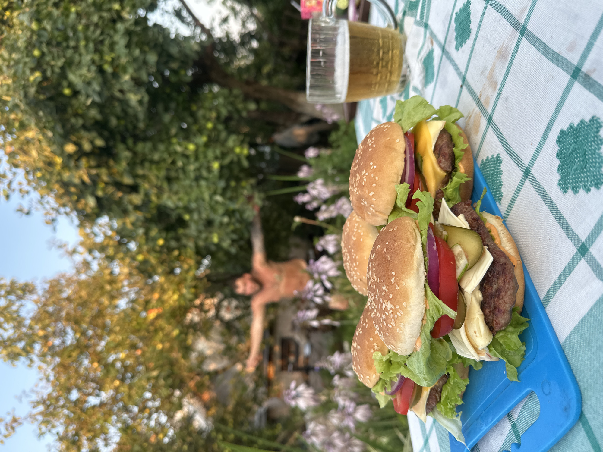 In the city of Galewo, Russia, a table outdoors is set with four cheeseburgers and a glass of beer. The burgers are arranged on a blue cutting board and are topped with lettuce, tomato, onion, cheese, and pickles. The table is covered with a green and white checkered tablecloth. In the background, a shirtless man with his arms outstretched stands in front of trees and purple flowers. The scene appears to be in a backyard or garden.
