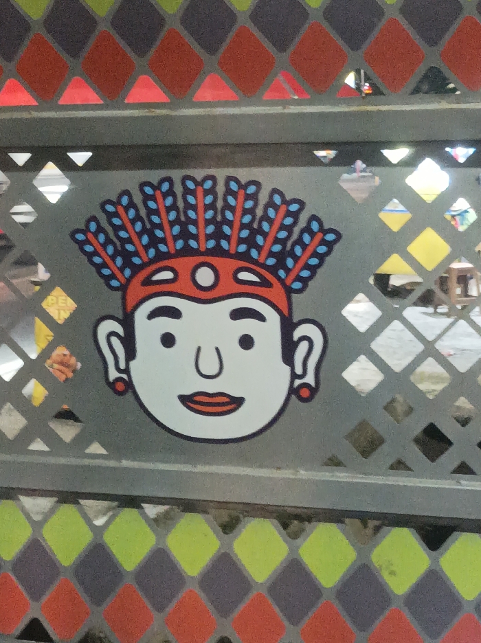 This image presents a close-up, frontal view of a stylized cartoon character affixed to a grey metal barrier or fence. The character features a white face, black dot eyes, a U-shaped nose, red lips, and two small red circular earrings. A prominent red headpiece adorned with multiple blue, feather-like elements outlined in red sits atop its head. This artistic rendition is consistent with traditional Betawi cultural motifs, such as "Ondel-ondel" or "Jali-Jali" figures, thereby situating the scene within the Special Capital Region of Jakarta, Indonesia.
The character is centered on a solid grey section of the barrier. Above this section, the grey metal is perforated with a grid of diamond-shaped cutouts, revealing a heavily blurred outdoor background. Vague shapes, including a partial yellow sign with dark lettering "PE..." on the left, and indistinct forms resembling wooden furniture, are discernible through the apertures. Below the character, the barrier transitions into a decorative pattern of solid, filled-in diamond shapes, colored in alternating red, dark grey, and lime green. No discernible individuals or explicit actions are present; the image focuses solely on the static decorative element and its immediate environment.