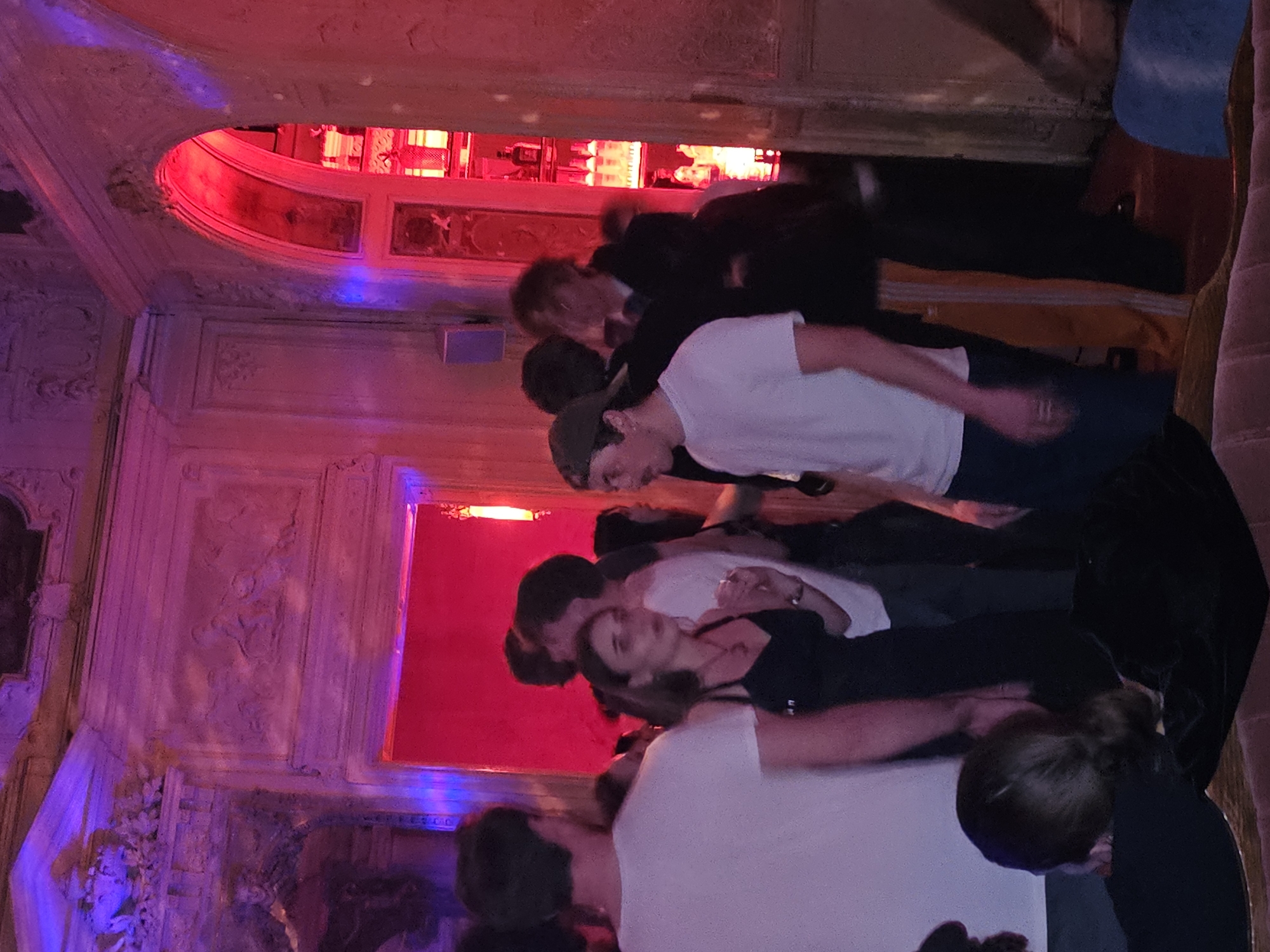 A crowded indoor scene in Paris, France features multiple people. The subjects are close together, some wearing white shirts, others in black attire. The background is dominated by ornate architectural details, with a vibrant red light source illuminating a window area, casting a contrasting purple hue on the surrounding walls. The people are engaged in some form of interaction, possibly a social gathering or event.