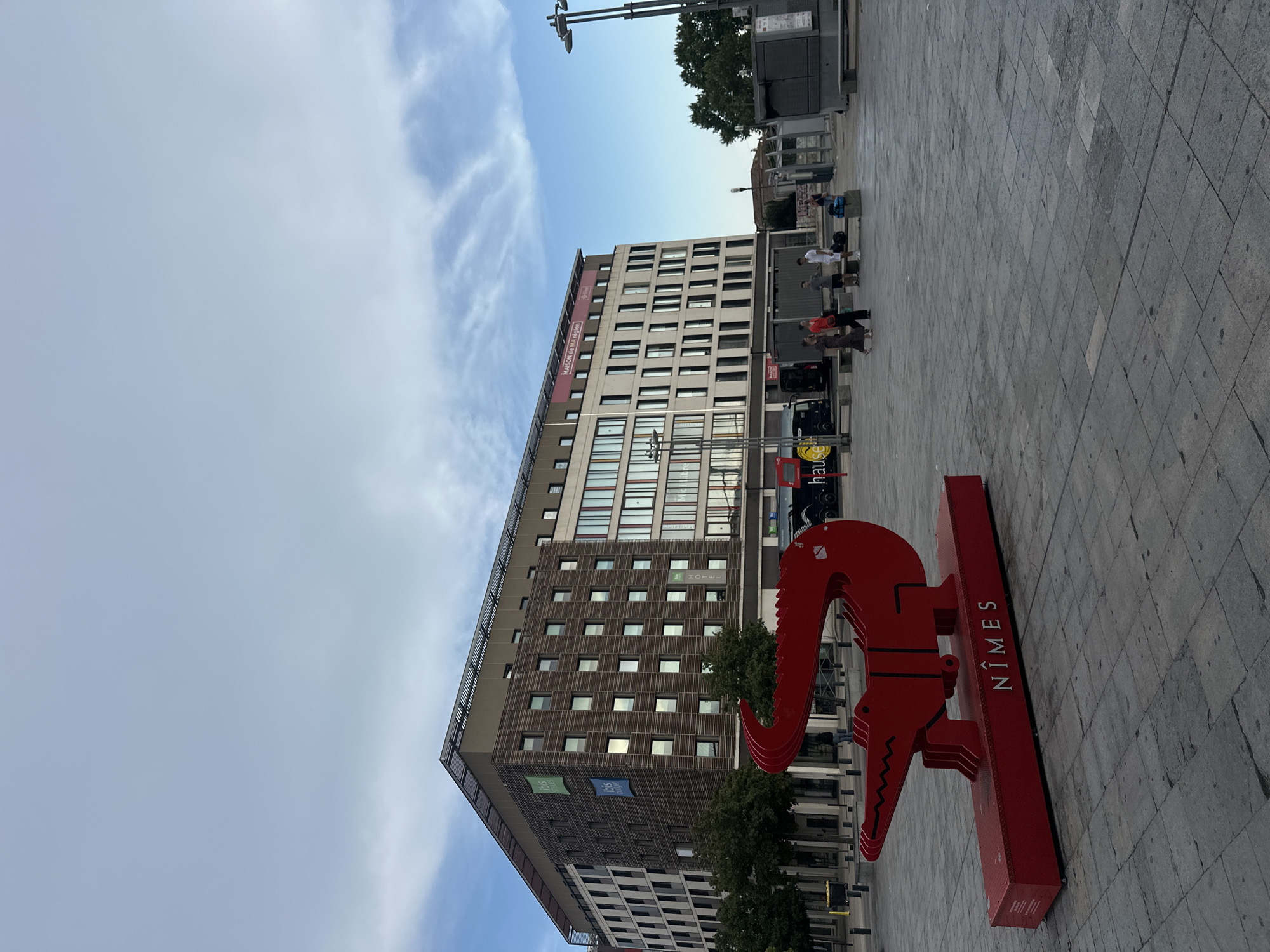 The media file displays an outdoor scene in Nîmes, France. In the foreground, a bright red sculpture resembling a stylized crocodile is prominently featured. The sculpture is positioned on a rectangular base, and text reading "NÎMES" is visible vertically along the side of the base.
Behind the sculpture, a modern, multi-story building with a beige facade and numerous windows dominates the background. Banners are visible on the building, with one clearly displaying the word "MAISON DE LA R...". Another banner shows the "ibis HOTEL" logo.
The ground is paved with large, grey square tiles, suggesting a public square or pedestrian area. Several people are scattered throughout the scene, walking or standing. They are dressed in casual clothing, indicating a daytime setting. The sky is partly cloudy, with streaks of white clouds against a blue background, suggesting fair weather. The lighting indicates it is likely daytime, possibly late afternoon or early evening, given the slightly soft light.
The overall impression is a public urban space with a striking art installation and a modern building. The presence of the "NÎMES" text on the sculpture and the "ibis HOTEL" on the building strongly suggest the location is Nîmes, France.