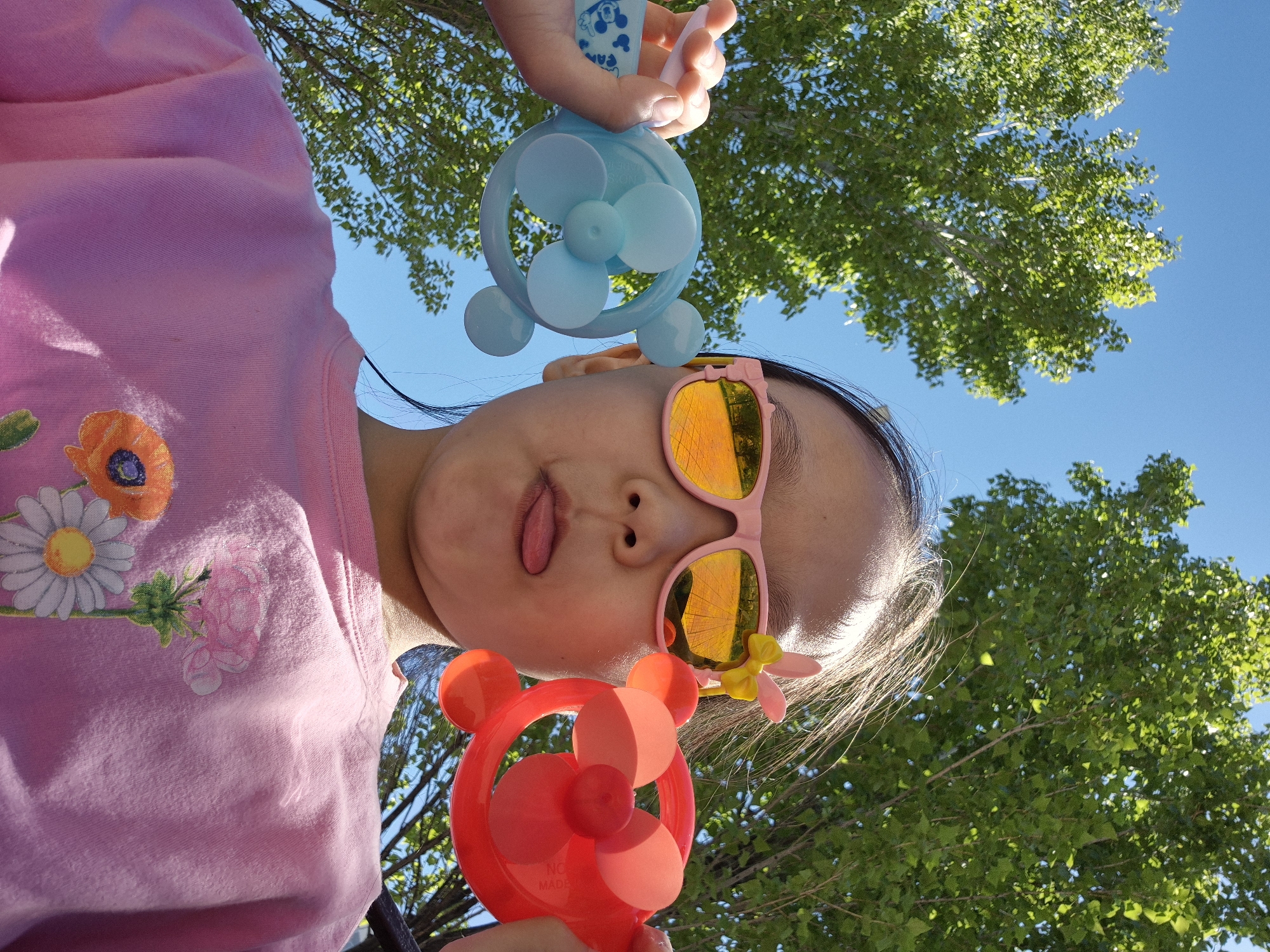 A young girl with dark hair and pink sunglasses is depicted in a bottom-up perspective, sticking her tongue out at the camera. She is wearing a light pink shirt with floral designs. Above her is a blue, hand-held fan being held by someone out of view and a red, hand-held fan. The background includes green trees and a clear blue sky. The location is Kokshetau, Kazakhstan.