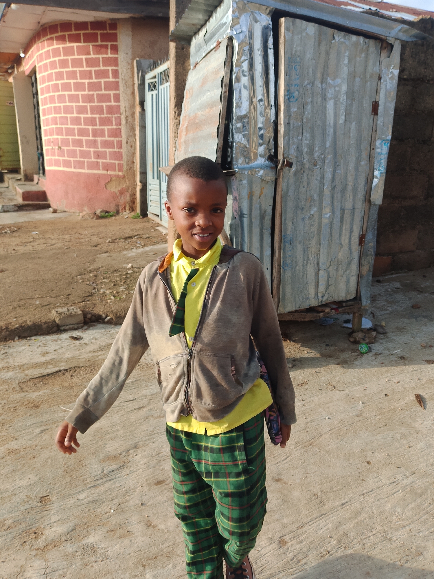 A young Black boy, approximately 7 years old, stands outdoors in a dusty, unpaved area that appears to be within a residential neighborhood in Jos, Nigeria. He is wearing a yellow collared shirt with a dark green and red plaid tie or scarf, a grey zip-up hooded jacket, and green and red plaid pants. His hair is short and dark. He is smiling and looking directly at the camera.
Behind the boy, there is a building constructed with red bricks and a curved facade on the left side of the image. To the right, there is a structure made of corrugated metal, possibly a makeshift dwelling or shop. A blue metal gate is visible in the background between the brick building and the corrugated metal structure. The ground is a mix of dirt and rough concrete. The overall impression is one of a developing urban environment, common in many parts of Jos.