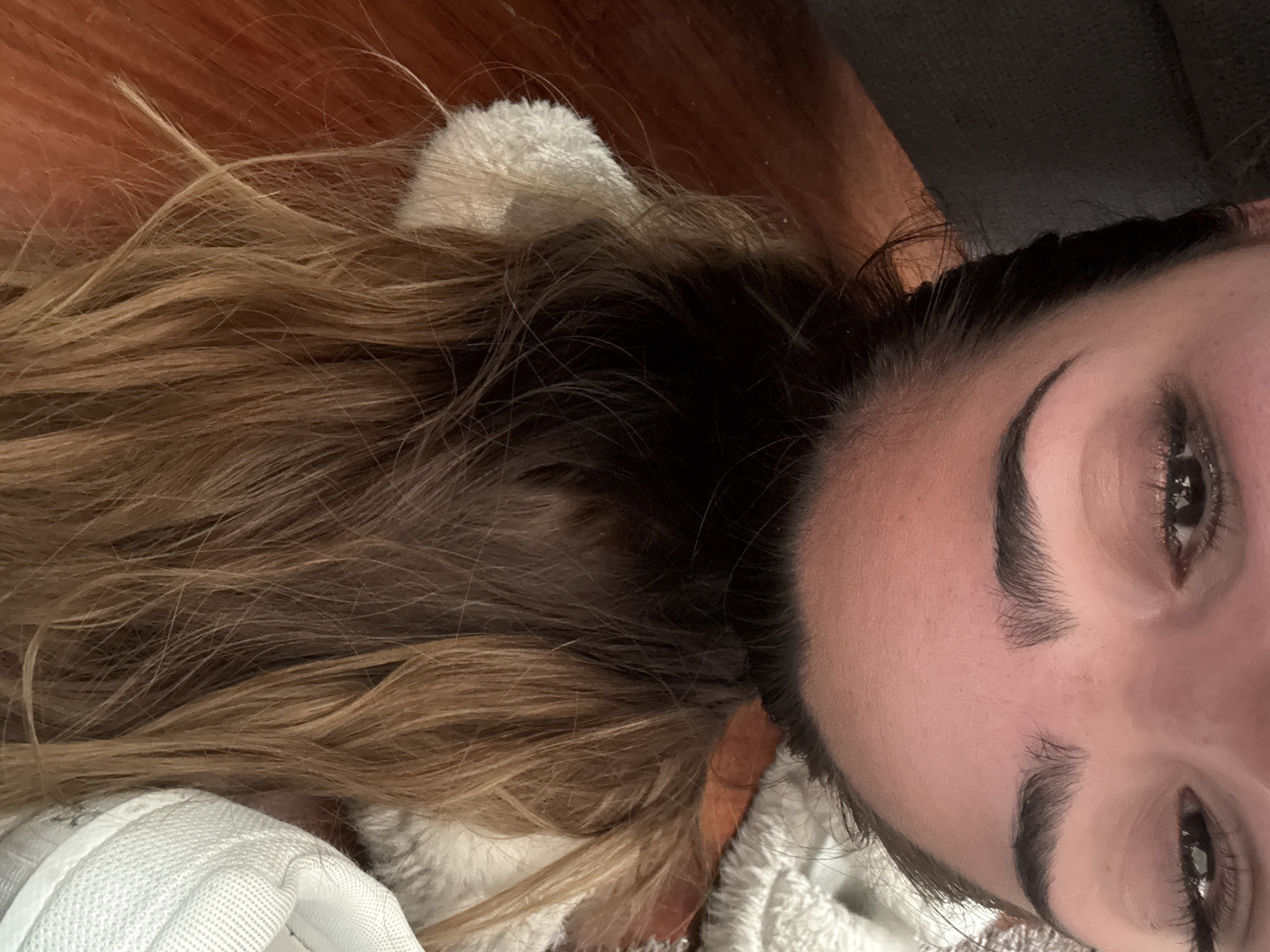 The image shows a person lying down with their head near the bottom right corner of the frame. The person has dark hair roots transitioning to blonde highlights. The person's face is visible, showing their eyes and eyebrows. A white, fluffy material is near the person's head and hair. The background appears to be a wooden surface. No location information is evident in the image.
