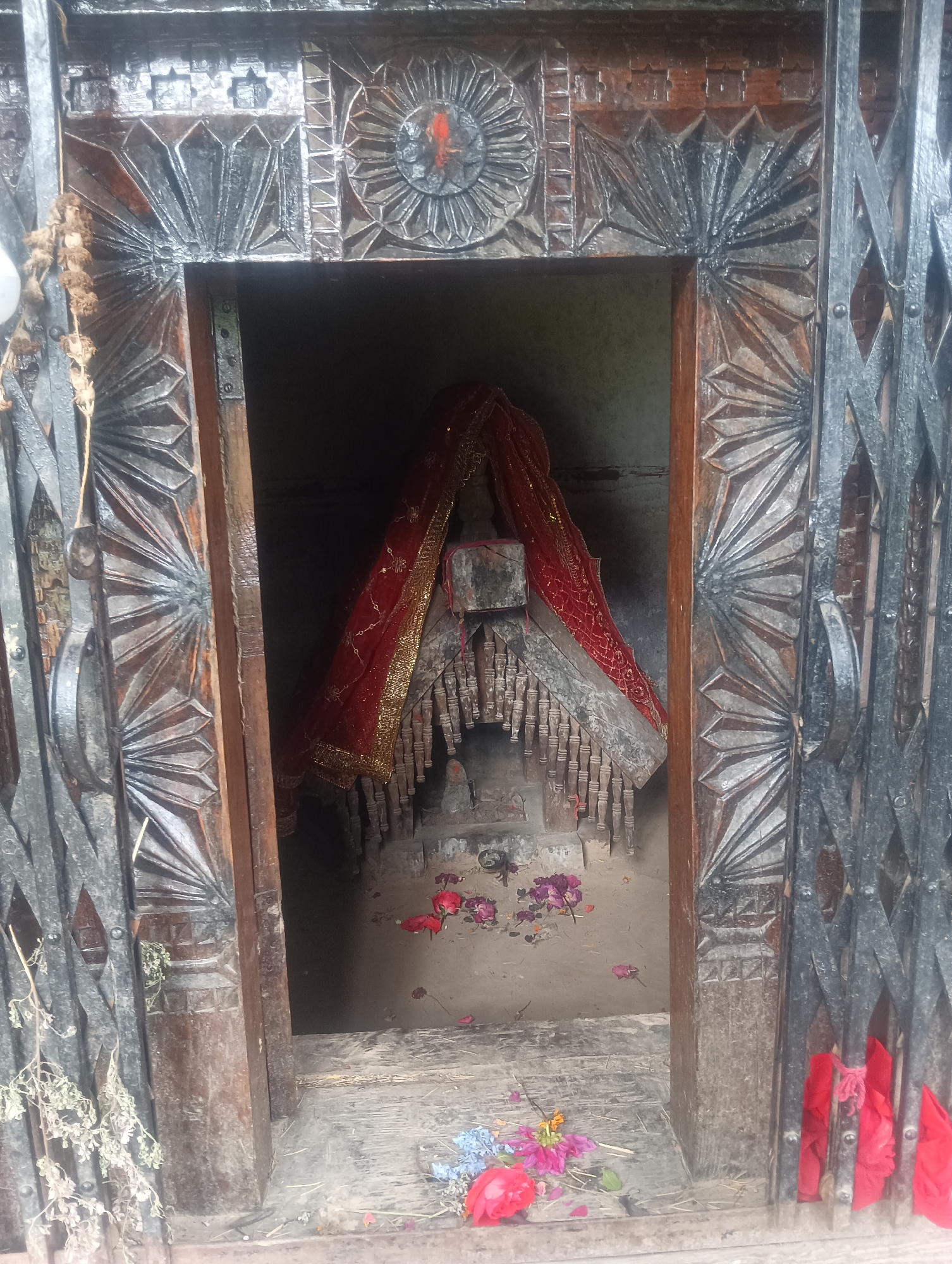This is a medium-shot, vertical, outdoor, eye-level, full-color photograph of an altar inside a doorway. The altar is adorned with a red cloth with gold trim, and there are scattered flowers in front of it. The doorway is framed by intricately carved dark wood and a black metal gate. The overall scene suggests a religious or spiritual site, likely a temple or shrine. The photograph appears to have been taken in Shanag, India, given the context provided.