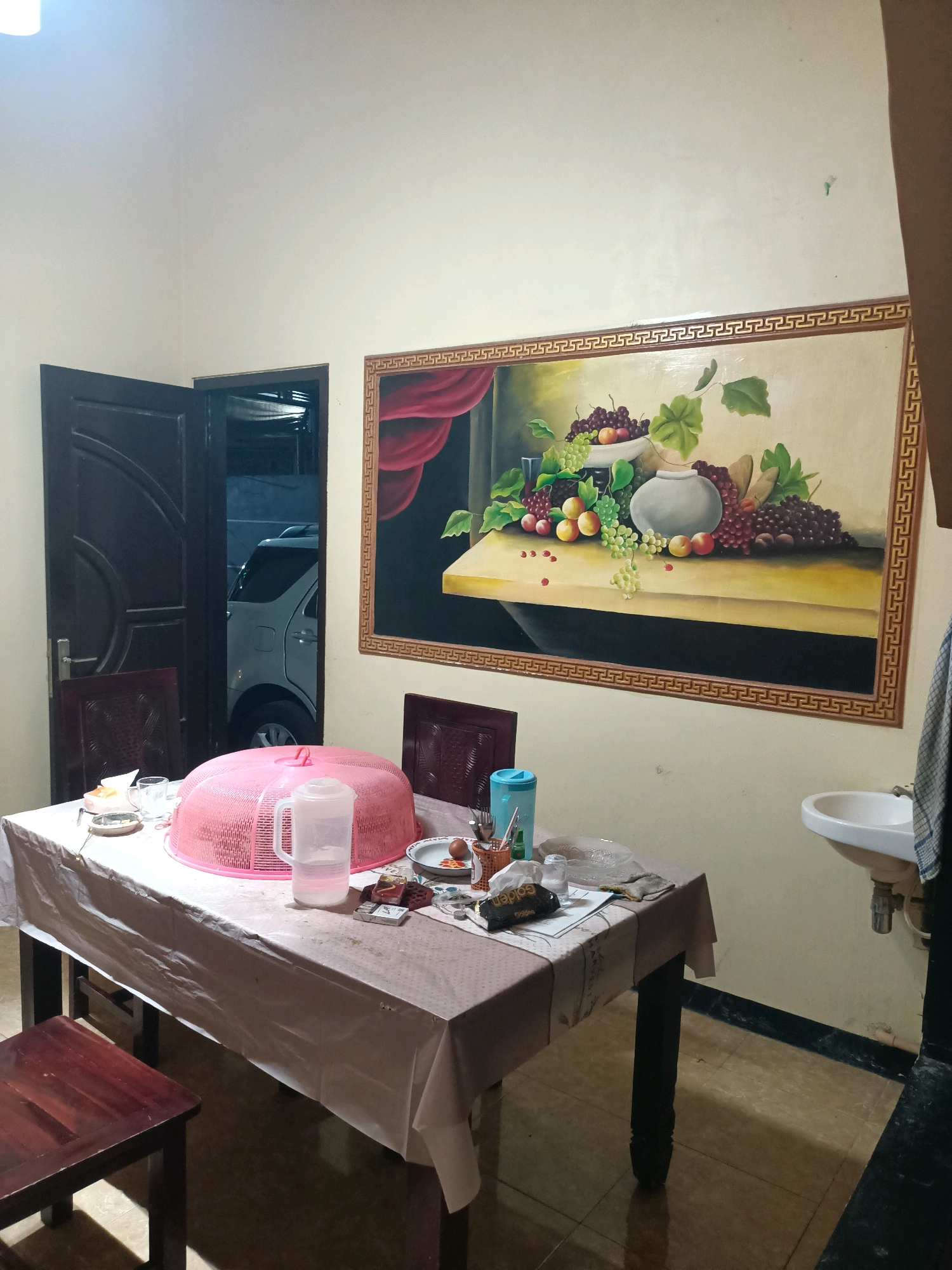 The image depicts an indoor dining area in Trenggalek, Indonesia. A dark wooden dining table is set with a patterned tablecloth and covered with a pink mesh food dome. On the table, there are several items including a plastic pitcher of water, a teacup and saucer, a pack of cigarettes, and a small plate with an egg. To the left of the table, a dark wooden chair is partially visible, and further left, a dark wooden door is ajar, revealing a glimpse of a white SUV parked outside. On the wall above the table, a large framed painting of a fruit still life, featuring grapes, apples, and a vase, is prominently displayed. To the right of the painting, a white sink is mounted on the wall. The room has light-colored walls and tiled flooring.