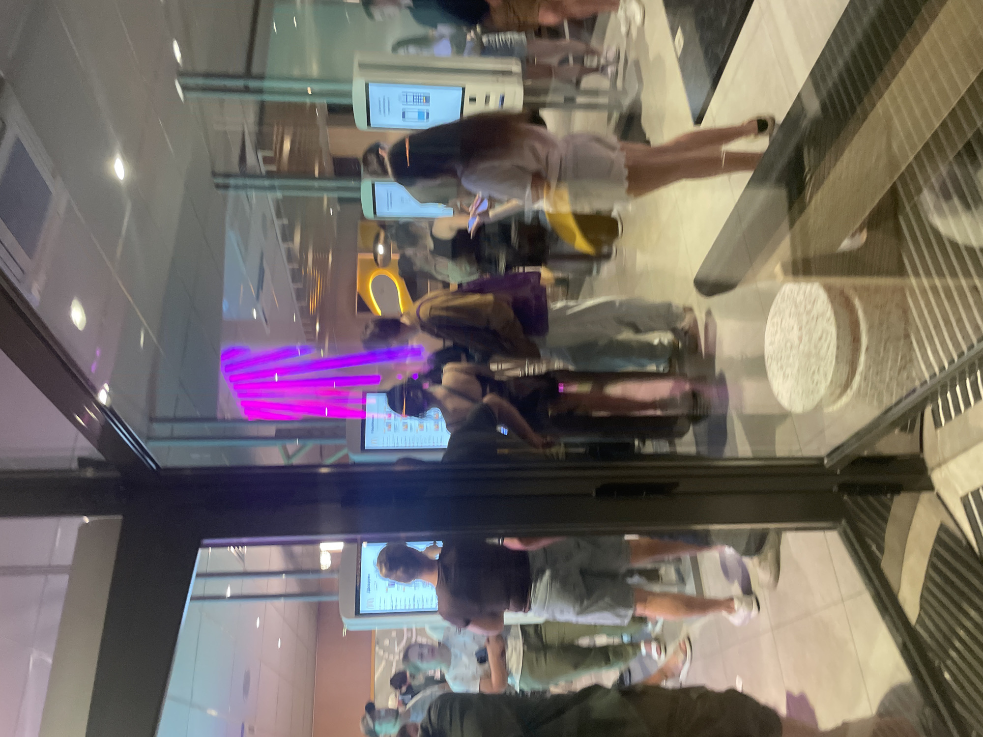 The image shows the interior of a fast-food restaurant, likely a McDonald's, as indicated by the digital menu boards and the overall ambiance. The scene is viewed through a glass storefront, with reflections present. Several people are visible, some standing in line and others interacting with the self-order kiosks. One person is looking at a digital menu board, another appears to be paying at a kiosk, and others are waiting. The lighting is bright, with a mix of overhead fluorescent lights and the glow from the digital screens. In the background, there are tall, bright pink light installations, possibly part of the restaurant's decor. The floor appears to be tiled. The setting suggests a busy evening or daytime service. The presence of McDonald's ordering kiosks and the general appearance of the establishment strongly suggest it is located in Ukraine, specifically Kyiv, given the prevalence of McDonald's outlets in the city.