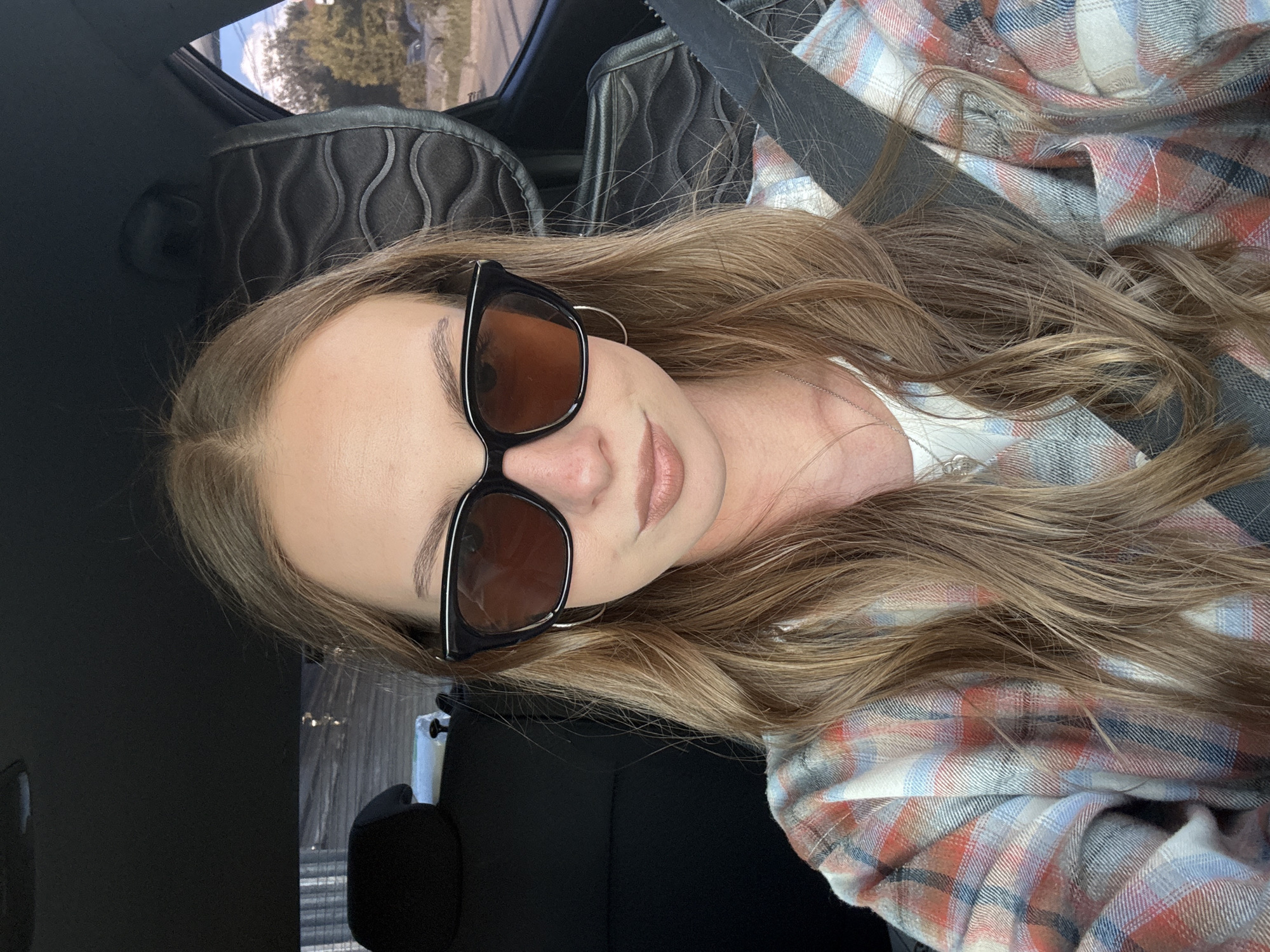 A young woman with long, blonde hair and sunglasses is taking a selfie in a car. She is wearing a plaid shirt and a seatbelt. The car appears to be parked outdoors, with trees and buildings visible through the window. The image does not contain enough information to determine the specific location in the city of Perm, Russia.