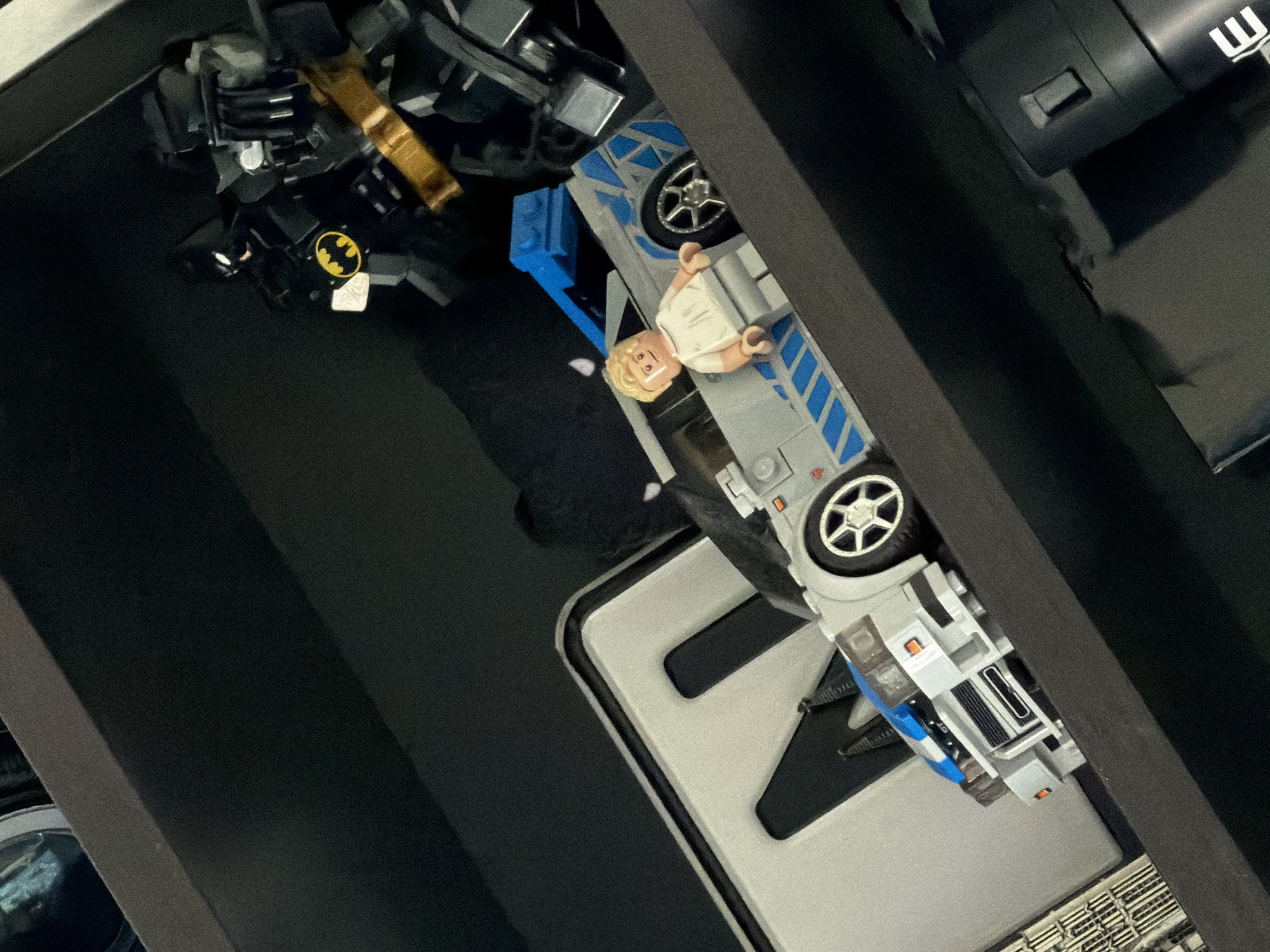 A Lego Batman figure stands behind a Lego car with a Lego figure seated inside. The Lego Batman is a dark, armored figure with a yellow bat symbol on its chest. The Lego car is white with blue accents and is positioned on a dark shelf. The Lego figure in the car has blond hair and is wearing a white shirt and gray pants. In the background, a large white object with cutouts is visible. The scene is dimly lit, suggesting an indoor setting.