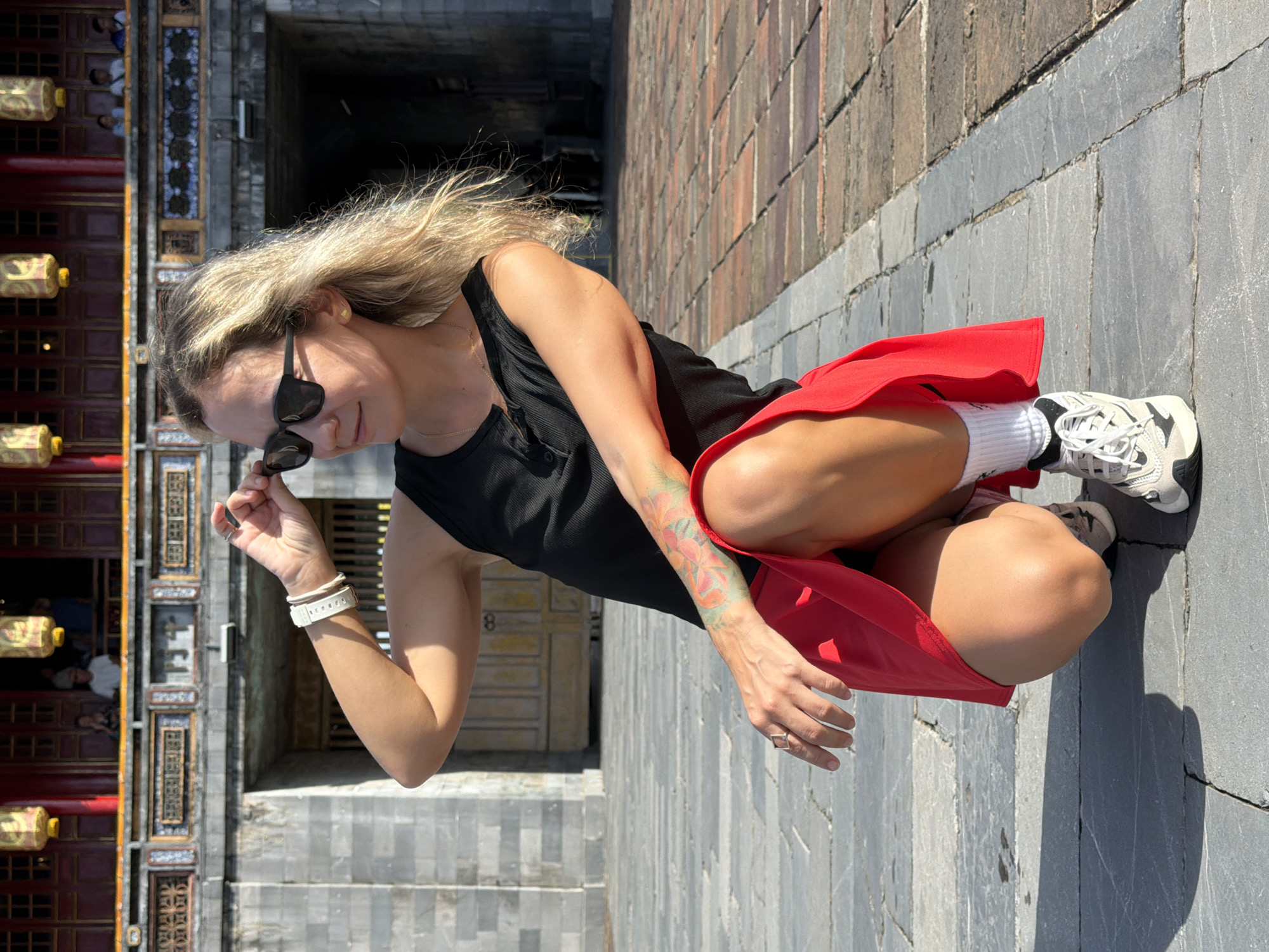 A woman with blonde hair, wearing sunglasses, a black tank top, and a red skirt, squats on a paved courtyard in front of a large stone structure. She is adjusting her sunglasses with her right hand. Her left hand rests on her left knee. She has a tattoo on her left arm. She is wearing white socks and athletic shoes. The stone structure behind her appears to be an ancient building with ornate decorations and what looks like a traditional Asian architectural style, possibly part of the Imperial City of Huế in Phú Xuân Ward, Vietnam, given the context. The courtyard is paved with large stone slabs. The lighting suggests it is daytime, with harsh shadows.