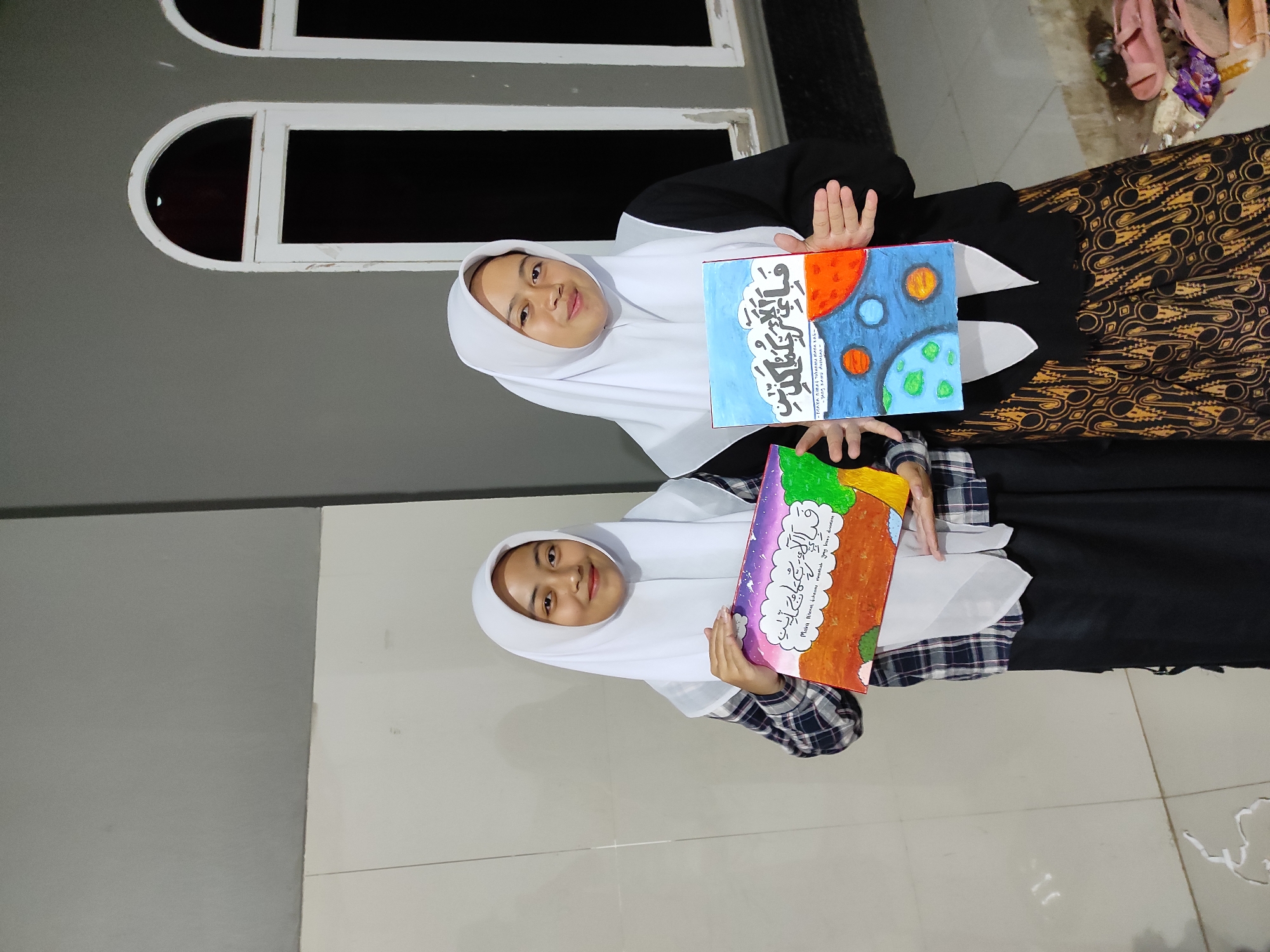 Two young women wearing white hijabs stand outdoors, holding up paintings. The woman on the left holds a painting with a dark, starry sky and a colorful landscape below. The woman on the right holds a painting depicting planets in space against a blue background, with Arabic calligraphy at the top and text in a smaller font below. They are standing in front of a building with a grey wall and arched windows. The building appears to be located in Sumedang, Indonesia, a city known for its natural beauty and cultural heritage, including traditional batik patterns often seen in local attire.