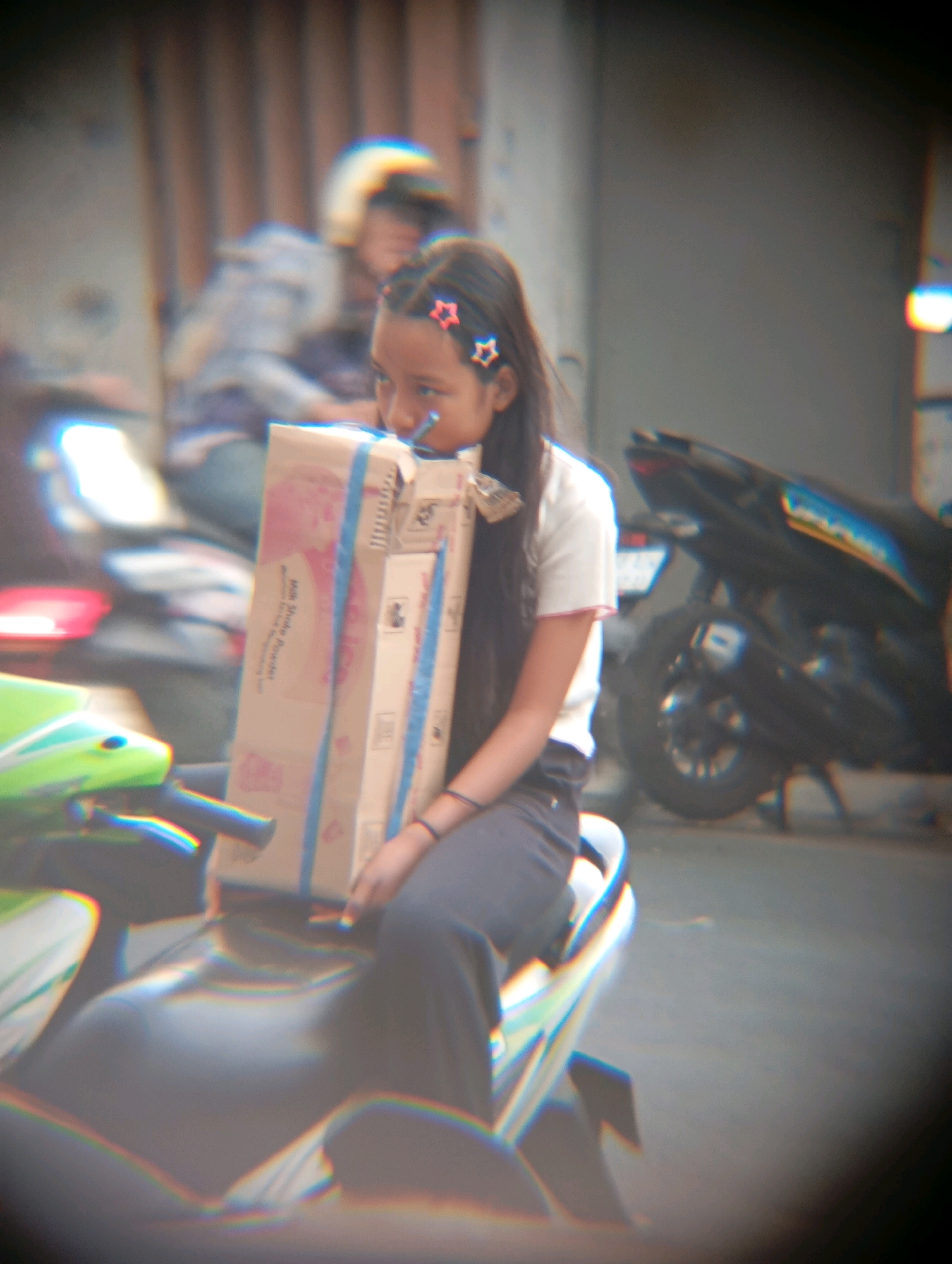 The image shows a young girl with dark hair, adorned with star-shaped clips, sitting on the seat of a scooter. She is holding a large, rectangular cardboard box, possibly for delivery or transport, which is secured with blue tape. The girl is wearing a white t-shirt with pink trim and dark pants. She is leaning forward, appearing to be either about to drink from or speak into a straw-like object protruding from the box.
Behind the girl, another person is partially visible, wearing a helmet. A black motorcycle is parked to the right of the girl, its rear wheel and exhaust pipe clearly visible. The background suggests an urban setting, possibly a street in Malang, Indonesia, characterized by muted colors and a somewhat blurred, weathered appearance, suggesting either the time of day or the environmental conditions. The overall scene is one of everyday activity, with a focus on the girl and the object she is holding.