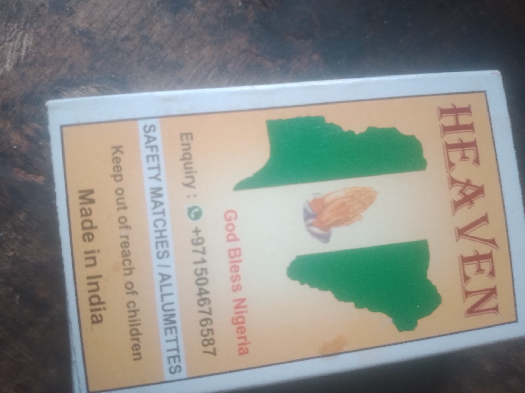 The image is a close-up of a matchbox with text and graphics. The matchbox is oriented vertically. The top portion of the matchbox has the word "HEAVEN" written vertically in a large, serif font. Below "HEAVEN" are two green shapes resembling landmasses, with a depiction of praying hands in between them. To the left of these graphics, there is text that reads "Enquiry: +971504676587", "God Bless Nigeria", "SAFETY MATCHES / ALLUMETTES", and "Keep out of reach of children". At the bottom left of the matchbox, it says "Made in India" vertically. The matchbox is placed on a dark, textured surface. There are no people visible in the image. The location is not explicitly mentioned in the image, however, "God Bless Nigeria" suggests a connection to Nigeria.