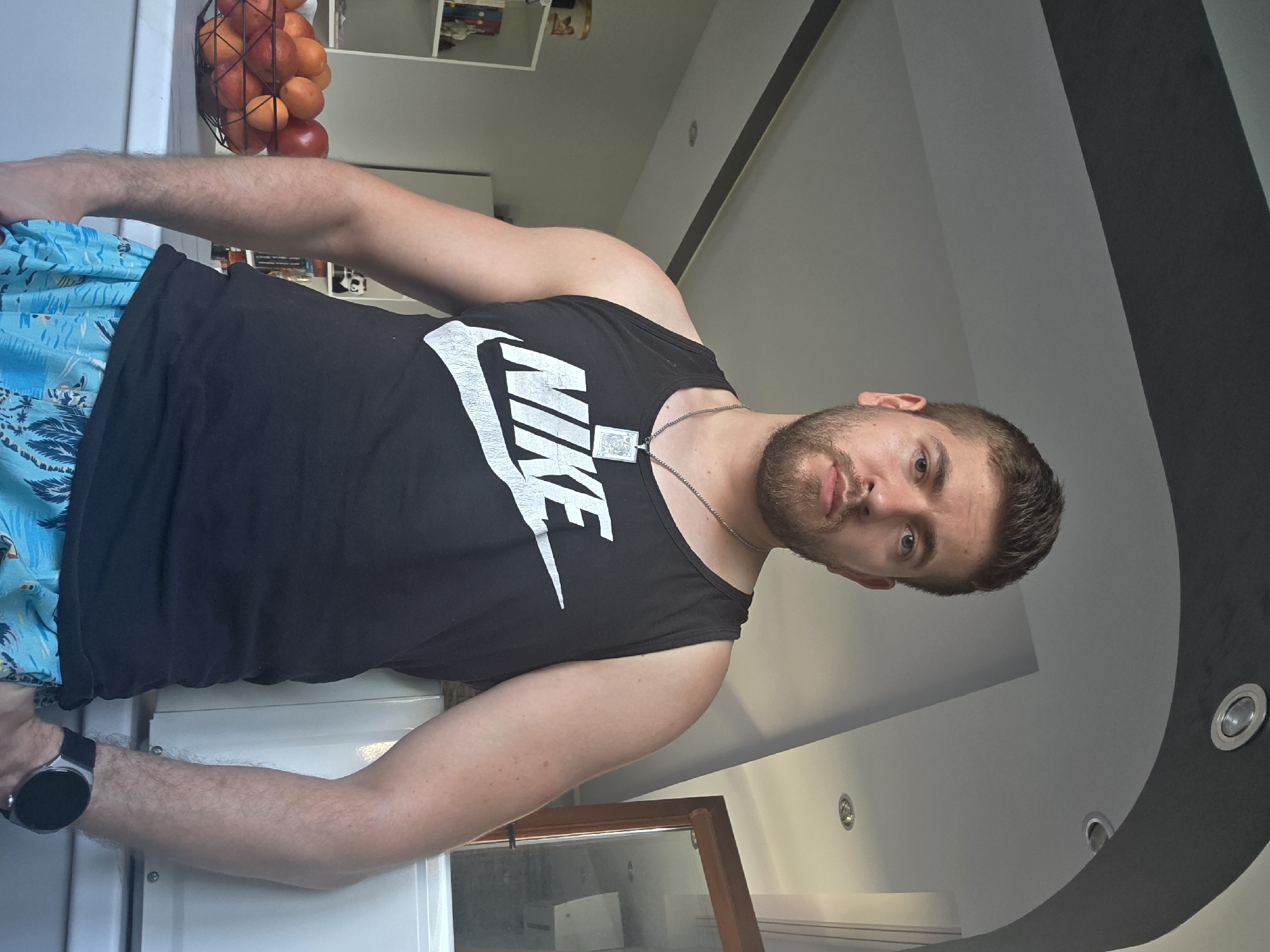 An adult male subject is depicted standing indoors, likely within a domestic setting in Yagodovo, Bulgaria. The subject has short, dark brown hair and a beard, light skin tone, and blue eyes, maintaining a neutral facial expression. He is wearing a black tank top with a white "NIKE" logo printed on the chest, light blue patterned shorts, a silver-colored chain necklace with a rectangular pendant, and a dark-colored smartwatch on his left wrist.
To the subject's left, a white countertop holds a black wire fruit bowl containing a variety of red and orange fruits, including pomegranates, apples, and oranges. Behind the countertop, a white shelving unit is visible, displaying several objects and books. To the subject's right, a white appliance is partially visible, alongside a wooden-framed opening, possibly a doorway or window, leading to another area. The ceiling is white with dark grey/black curved and straight architectural elements, incorporating recessed circular light fixtures. The walls are light-colored. The subject's hands are mostly obscured by his shorts and body.