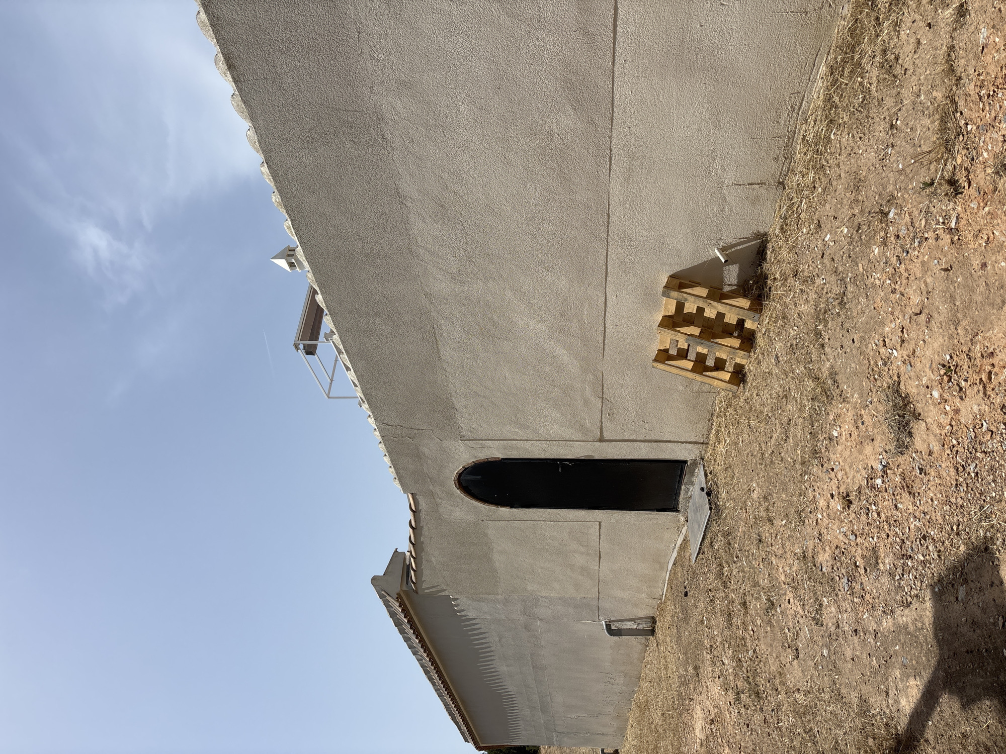 This is a photograph taken outdoors, likely in Orihuela, Spain, under a clear blue sky with some wispy clouds. The image is oriented vertically. The primary subject is the side of a light-colored building with a textured stucco finish. The building has a section of roof visible with terracotta tiles and a decorative, scalloped edge along the eaves.
To the left of the building, a portion of a white metal structure, possibly a balcony railing or scaffolding, is visible against the sky. On the right side of the building, there is a dark, arched doorway. Leaning against the wall near the doorway are two stacked wooden pallets. The ground in front of the building is covered with dry, light-brown grass and small stones, suggesting a dry climate or season. The overall impression is one of an exterior view of a building, possibly under construction or renovation, in a somewhat arid location. The lighting indicates it is daytime, likely mid-morning or afternoon, given the clear sky and the shadows cast. There is no visible text in the image.