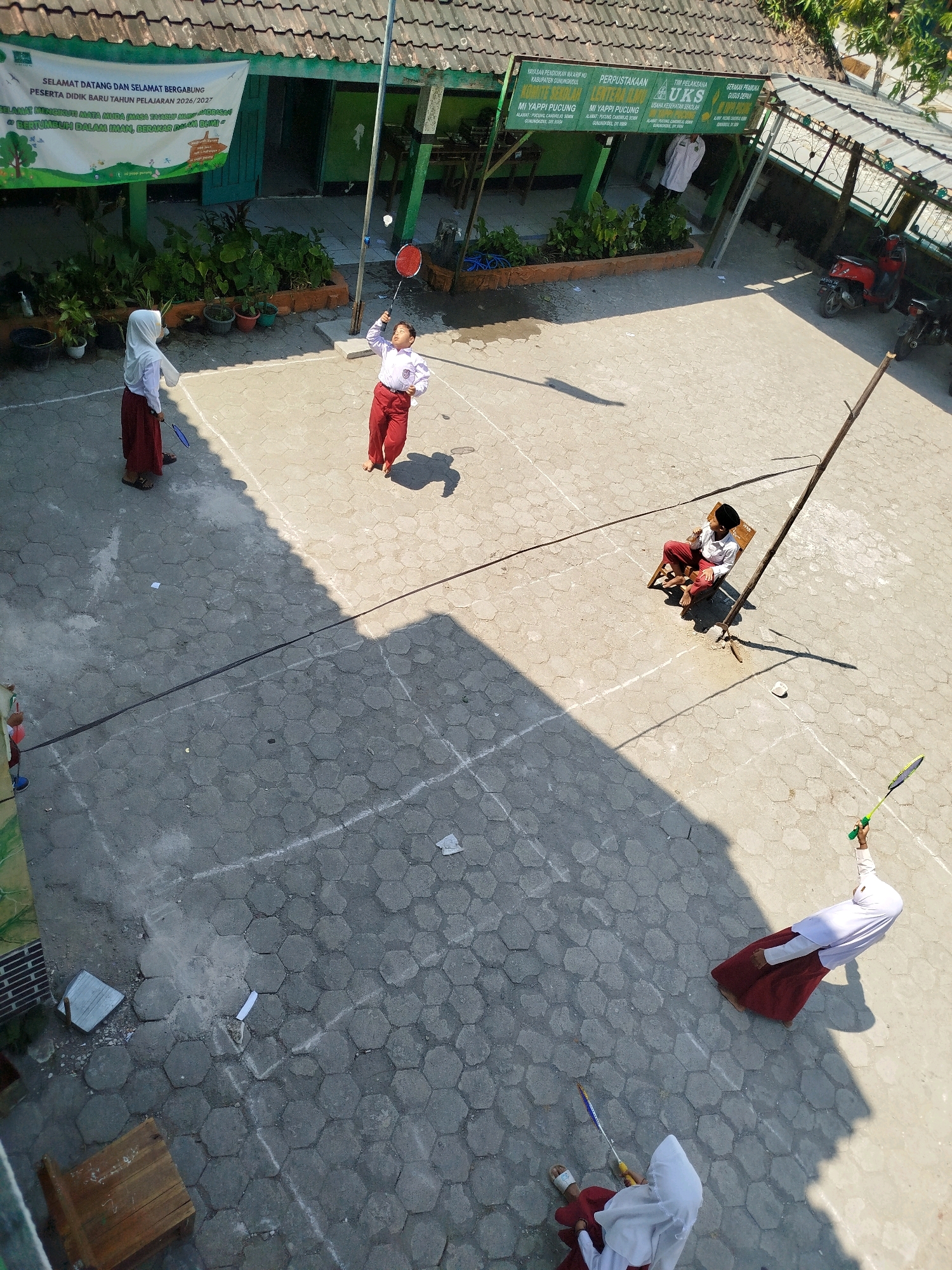 The image captures an overhead view of an outdoor courtyard area at what appears to be a school in Semin, Indonesia. The courtyard is paved with hexagonal concrete tiles. In the foreground, two students in white shirts and maroon skirts, wearing headscarves, are playing badminton. One student is on the left, facing away from the camera, holding a blue badminton racket. The other student is in the middle, facing left, mid-swing with a red badminton racket. Further back, a student sits on a wooden chair, watching the game. To the right, another student in a white top and maroon skirt is positioned as if playing badminton, holding a green racket. A tall, thin wooden pole stands in the center of the courtyard, casting a shadow. In the background, a building with a green facade and a tiled roof is visible, adorned with banners. One banner, with green and white text, welcomes new students. Other signs are also visible on the building, one of which appears to be for a "UKS" (Usaha Kesehatan Sekolah, School Health Unit). A red scooter is parked to the right of the building. The overall scene suggests a lively school activity day.