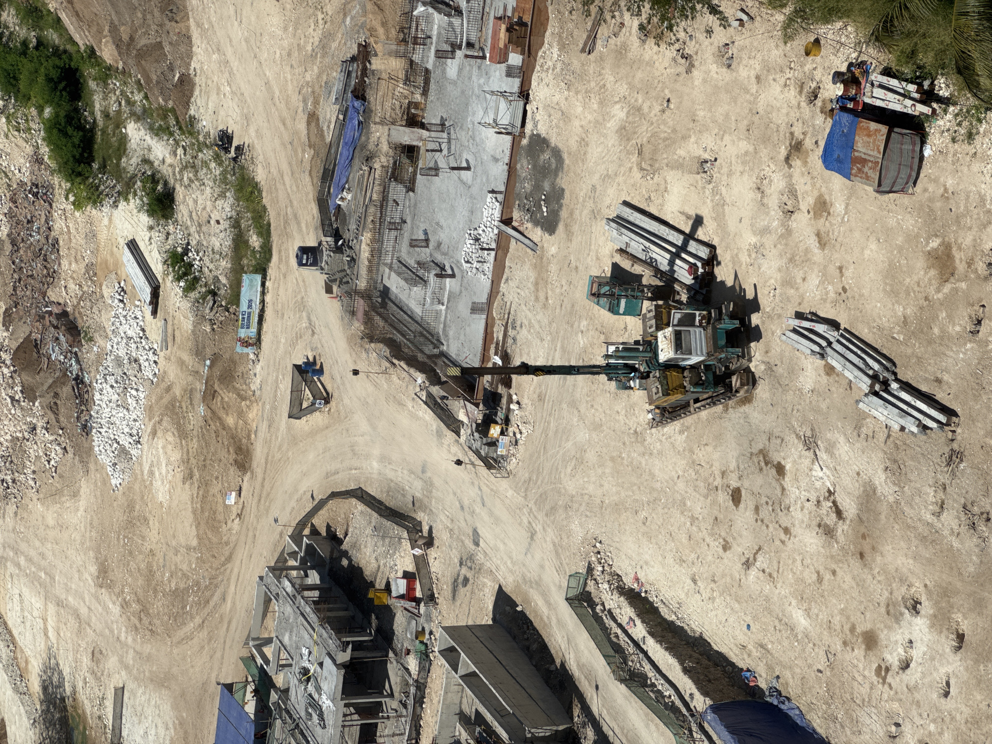 The image depicts an aerial view of a construction site in Kuta Selatan, Indonesia. A large crane is centrally positioned, with multiple stacks of concrete beams and materials scattered around the site. Partially constructed concrete structures are visible in multiple locations, including at the bottom of the image. The ground is mostly dirt and sand, with pathways and tire tracks indicating movement of vehicles and machinery. Some patches of vegetation are visible on the perimeter of the construction area. A small makeshift shelter or worker's camp, covered with a blue tarp, is situated on one side. Heavy machinery, likely used for excavation and material handling, is present throughout the site. There are a few people on the construction site but they appear to be spread out.