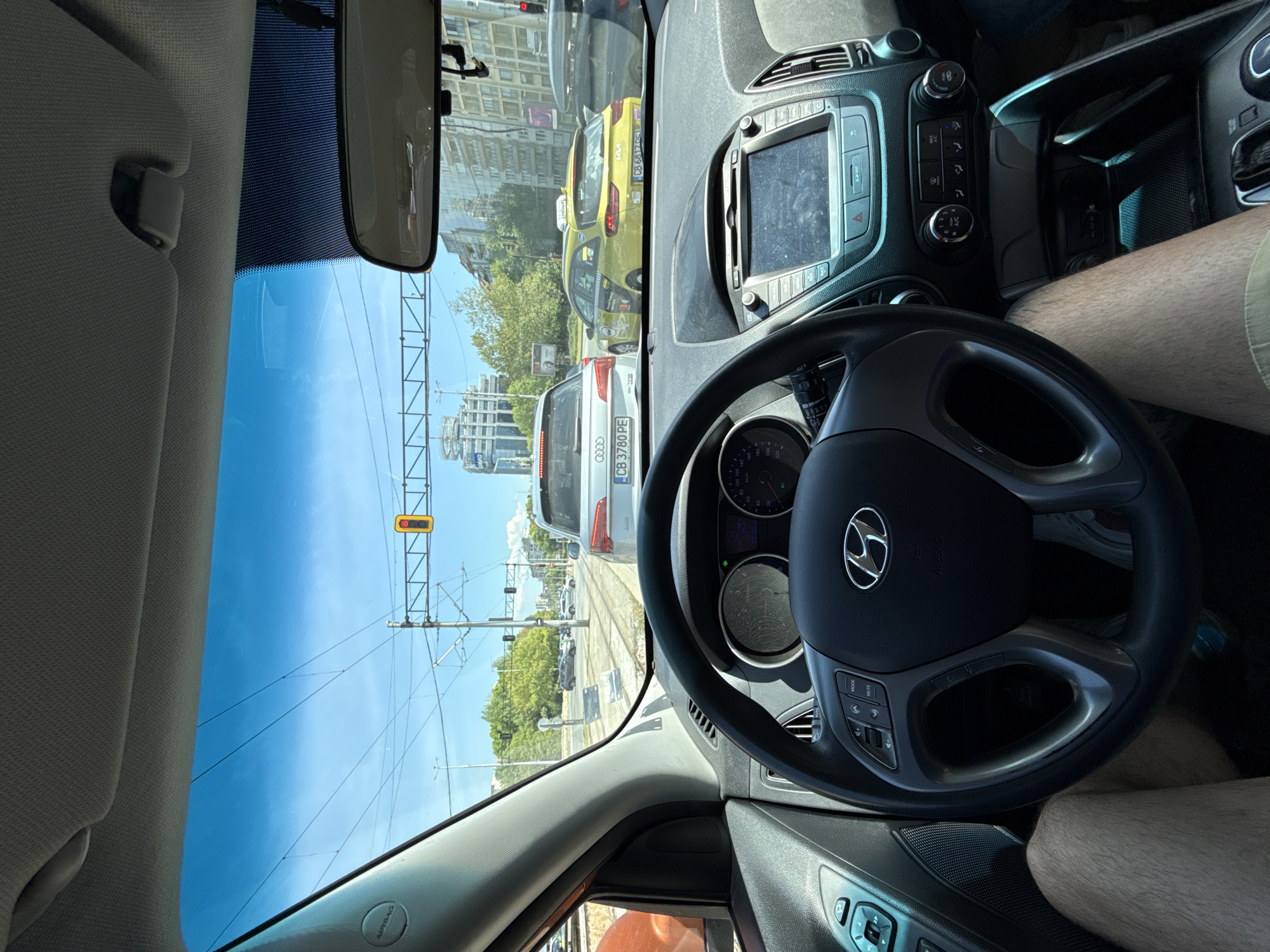 The view from inside a Hyundai car stuck at a tram crossing, facing forward. Outside, there's an Audi with license plate CB 3780 PE, a yellow taxi cab with license plate CB 6817 KB, buildings, a tram line overhead, and the sky. The tram crossing signal is red. The license plates on both cars indicate that they are registered in Sofia, Bulgaria.