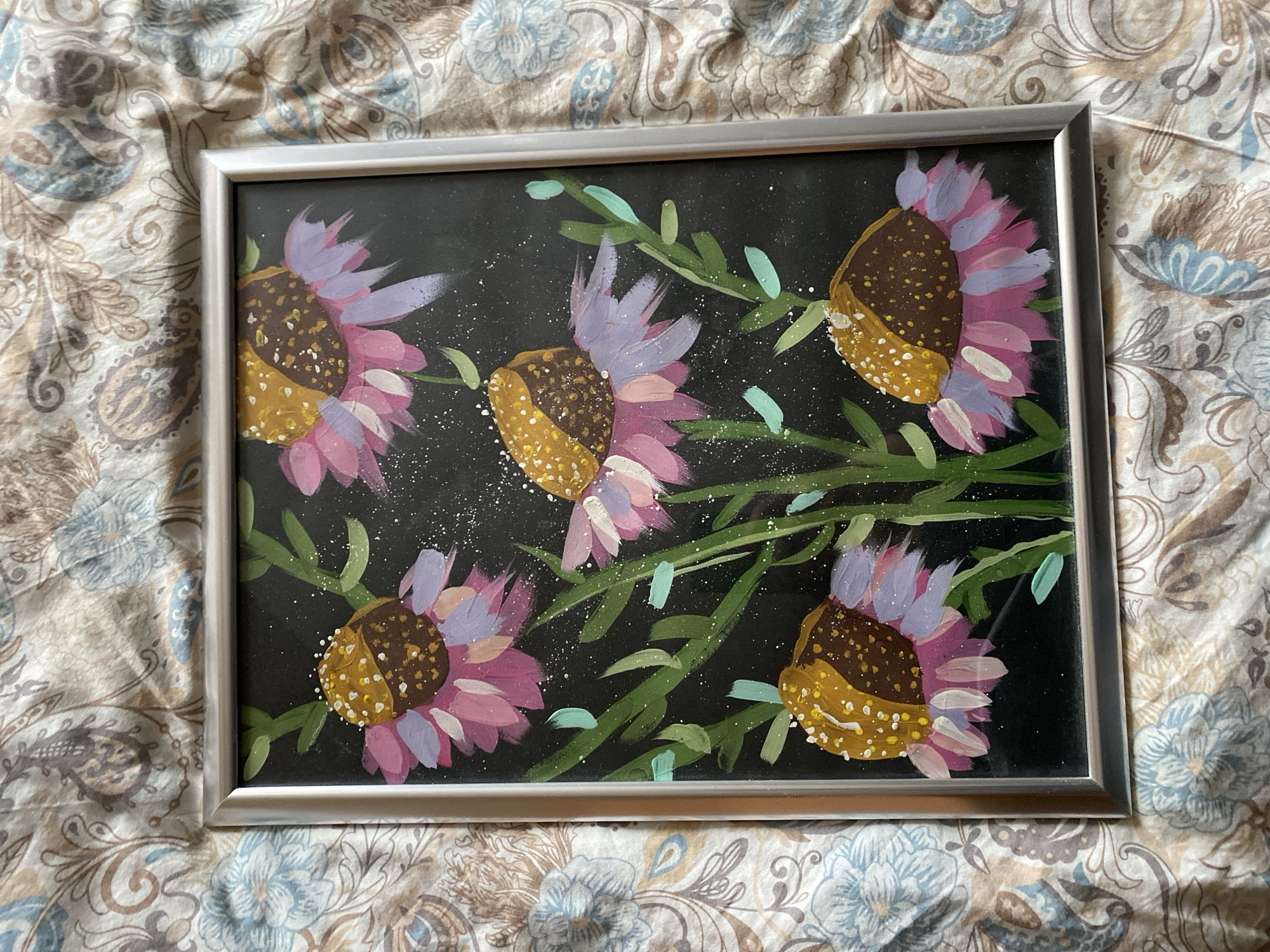 A framed painting of stylized sunflowers with a dark background is set against a patterned bedspread. The frame is silver-colored. The sunflowers have dark brown and yellow centers, pink and lavender petals, and green stems with small green leaves. The bedspread has a light background with a repeating floral and paisley pattern in shades of brown and blue. The scene is indoors. There is no information to suggest the city of Izhevsk, Russia.