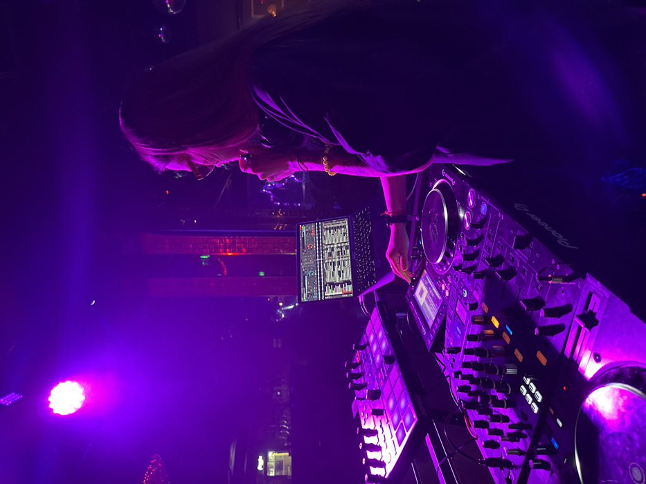 A DJ with long hair, glasses, and a black top works at a Pioneer DJ setup, including a laptop displaying audio waveforms, illuminated with purple stage lights. The scene is likely a club or performance venue, with ambient background lights and a dance floor visible. The DJ's hand is near the equipment, suggesting active mixing or track selection. The overall atmosphere is vibrant and energetic.