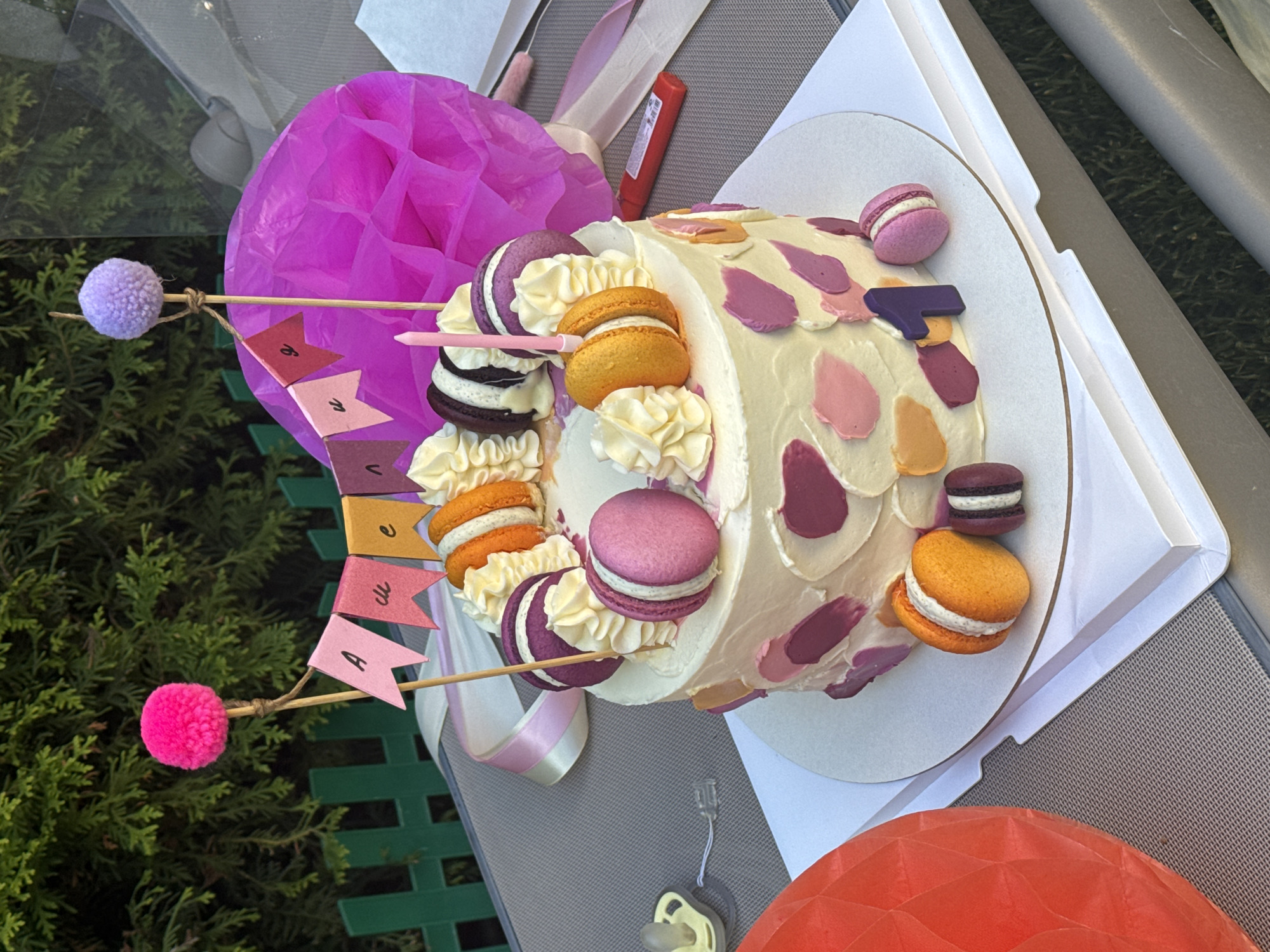 A brightly decorated birthday cake sits outdoors on a table. The cake is white with colorful splotches of pink, orange, and purple. It is adorned with macarons in pink, orange, and purple, as well as piped white frosting. A pink candle and a purple number "1" topper are also on the cake. A banner spelling out "Айжан" (Aizhan) in pink and orange flags hangs from bamboo skewers topped with pom-poms. In the background, there is a pink tissue paper pom-pom and lush green foliage, suggesting an outdoor setting. The foreground shows a portion of a red textured sphere and a pacifier. The image is likely taken in Sary-Oy, Kyrgyzstan, given the context of the user's request. The lighting suggests it is daytime.