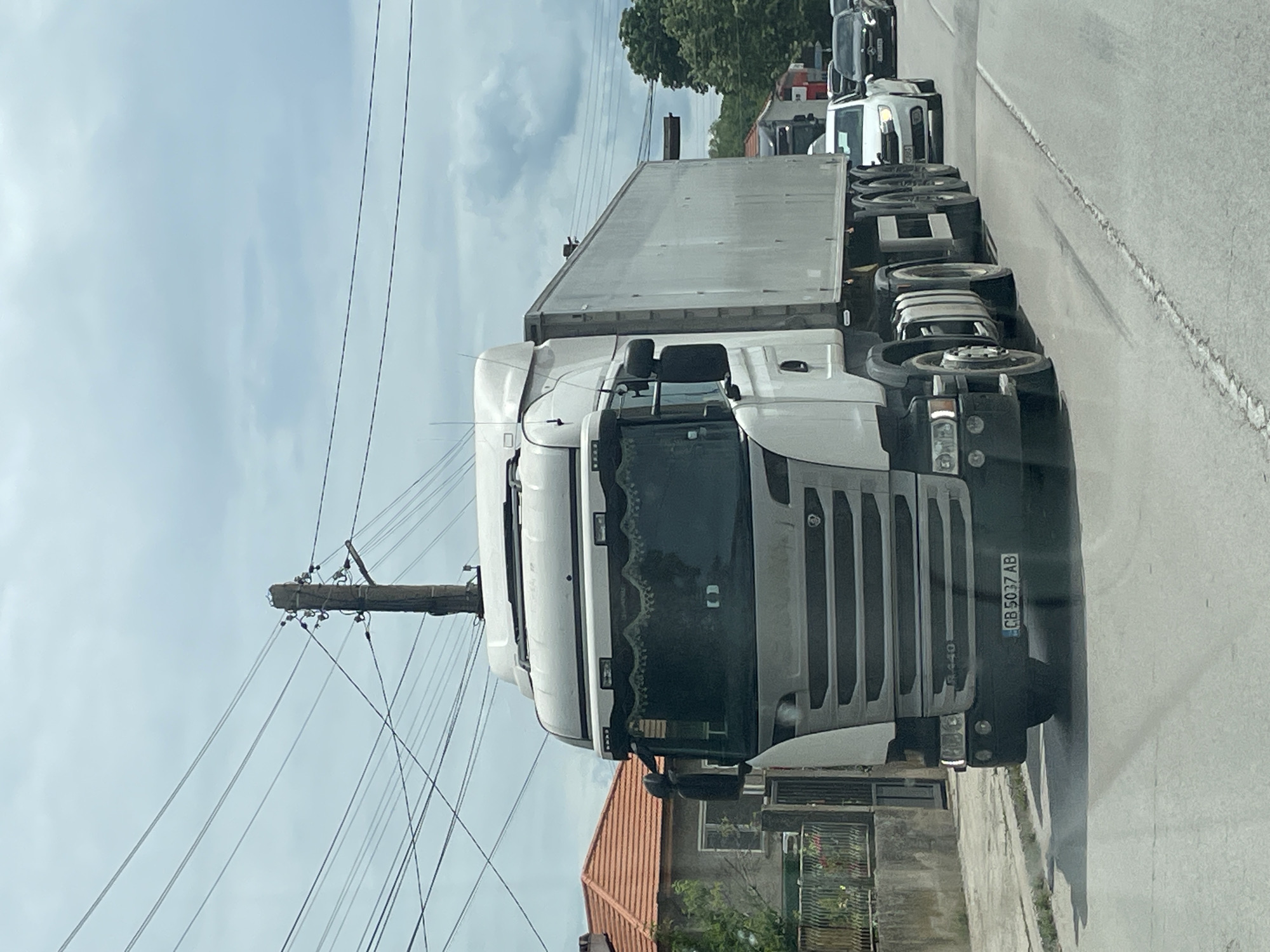 A medium shot of a large, white Scania R440 truck traveling on a road. The license plate reads "CB 5037 AB". The truck has a box trailer attached. Overhead electrical wires and a utility pole are visible. A low building with an orange tile roof and a fenced yard is to the left of the truck. Additional vehicles are stopped ahead of the truck in the distance. The image appears to be taken in Tsar Kaloyan, Bulgaria.