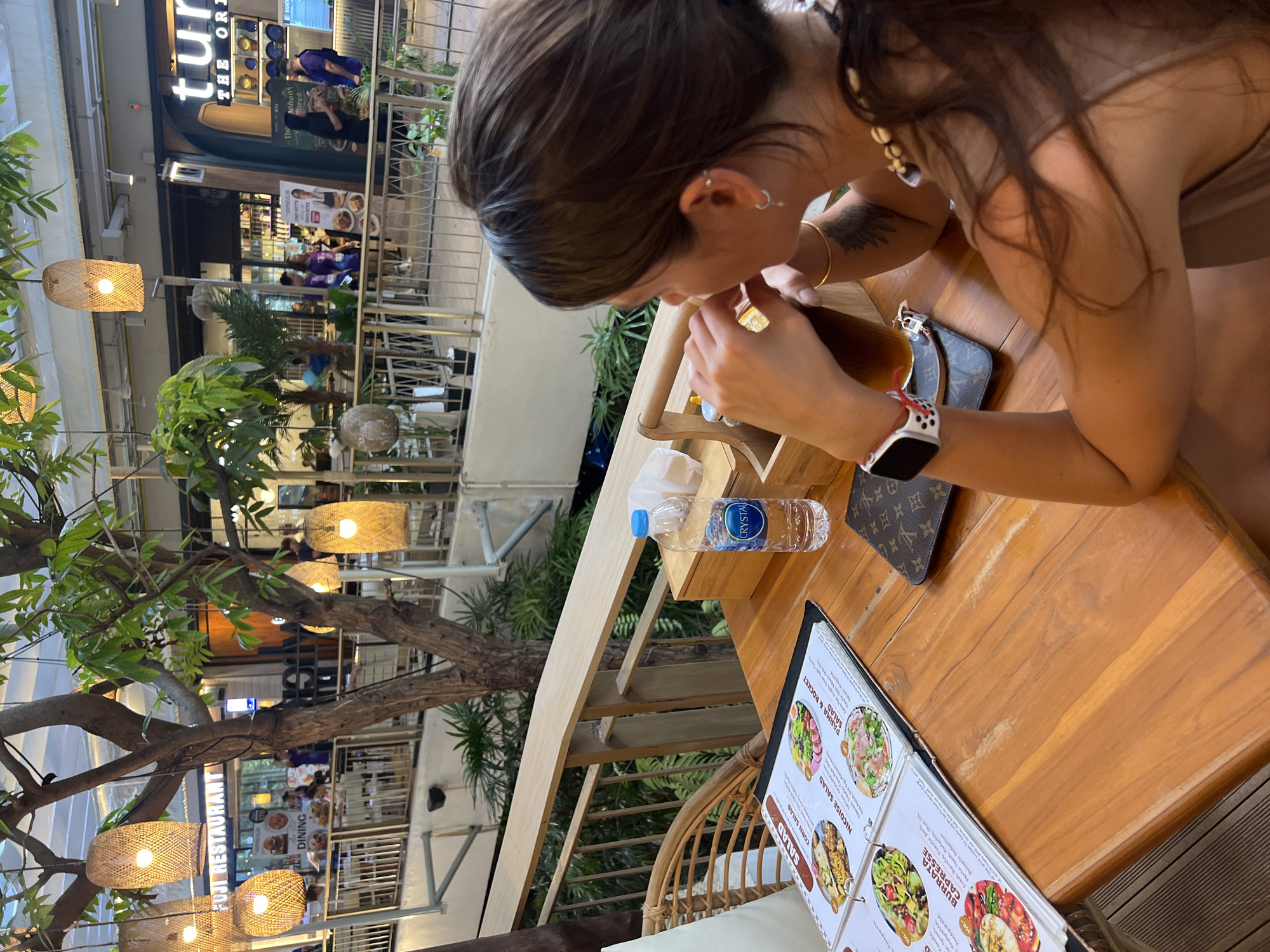 A medium, angled shot depicts a woman at a wooden table in an outdoor dining area, possibly in Unknown, Unknown. The woman, seen mostly from the side and back, has dark hair, tan skin, and multiple piercings in her ear. She wears a bracelet, a watch with a red and white band, and a light-colored top. She is drinking from a glass with a straw. Beside the glass is a Louis Vuitton wallet, a bottle of Crystal water, and a menu. The menu displays various salad options.
Behind the woman, the scene opens to reveal an outdoor restaurant setting with plants and woven light fixtures hanging from trees. In the background, the storefronts for "tur The Ori" and "FUJI RESTAURANT" are visible, indicating a commercial area. People are seen walking in the background, suggesting a bustling environment.