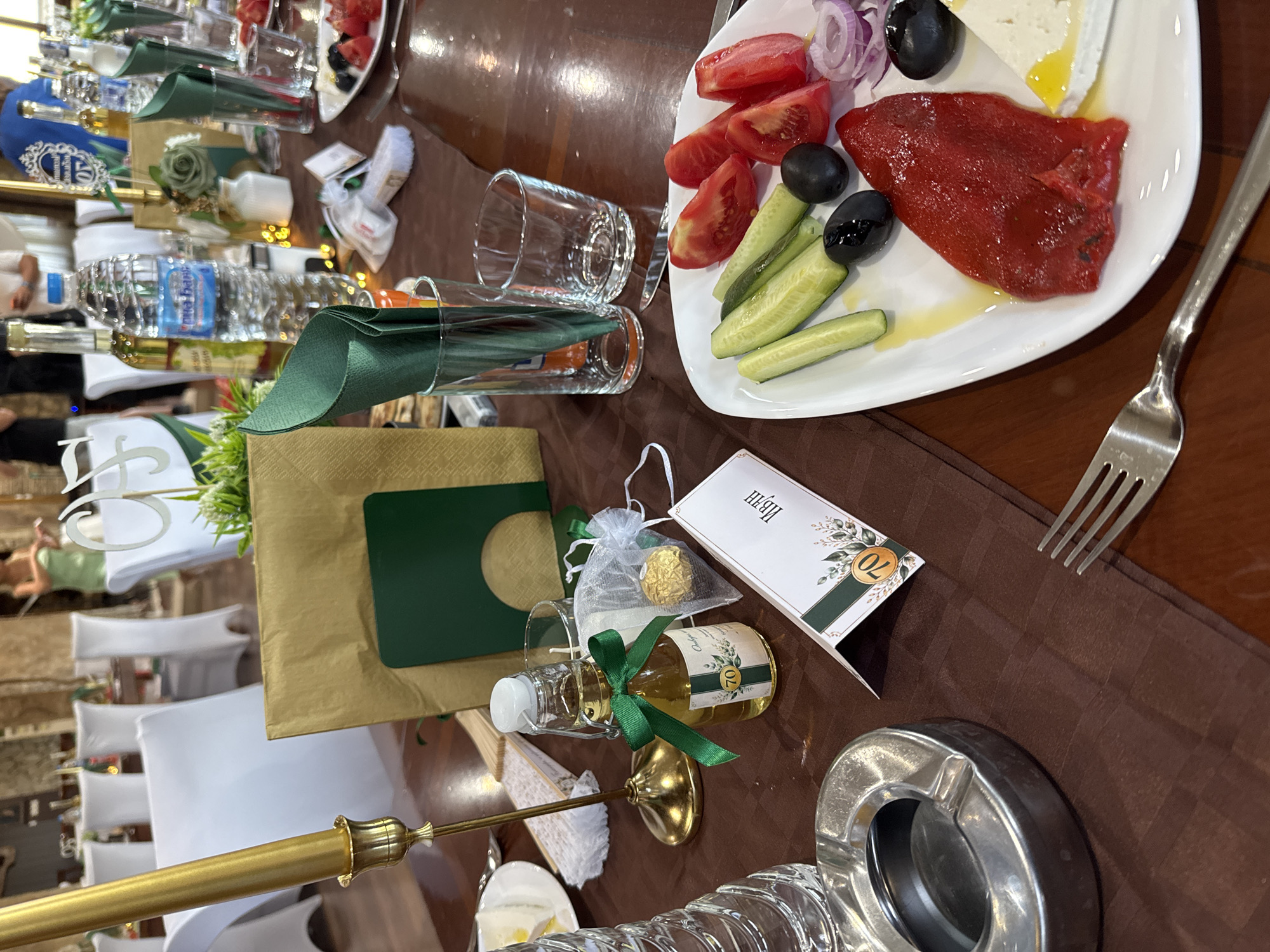 This is a photograph of a table setting at an outdoor event, possibly a celebration or reception, in Sredets, Bulgaria. The table is covered with a brown tablecloth. On the table are various items, including a gold candle holder with a gold candle, a small glass bottle with a green ribbon and a label that reads "70", a white gift bag with a gold chocolate, a white folded card with a green border and a floral design, green napkins, and several glasses. A white plate with a salad containing tomatoes, cucumbers, olives, red onion, and a piece of red pepper and white cheese is also visible. In the background, there are white-covered chairs and people dressed in formal attire. The overall impression is one of a festive gathering.