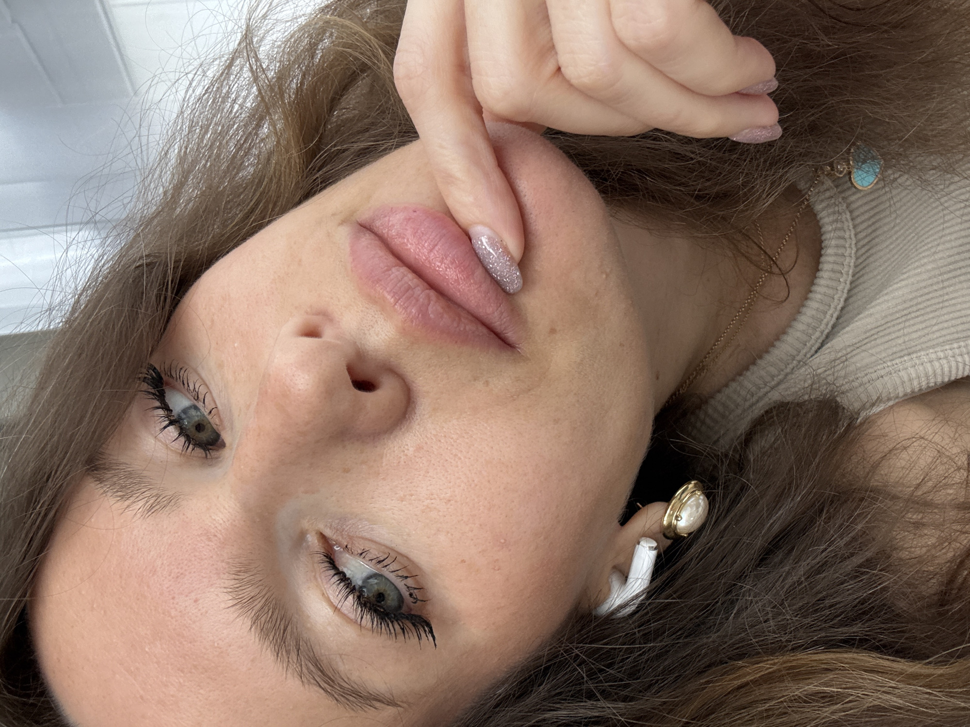 A close-up, high-angle shot captures a person lying down, looking towards the camera. The subject is a young adult with fair skin, brown hair, and blue-green eyes enhanced with dark mascara on long lashes. Their expression is calm and neutral.
The person's head is tilted, with their face occupying the left side of the frame. Their right hand is positioned near their mouth, with a finger gently touching their glossy, light pink lips, which appear to have been recently treated (e.g., lip blush or filler). The nails on this hand are manicured with a shimmering, glittery light pink polish.
The person is wearing a light-colored, possibly beige or cream, ribbed knit top. A thin gold chain necklace with a small, light blue, heart-shaped or teardrop-shaped pendant is visible around their neck, partially obscured by their hair. A white wireless earbud is visible in their right ear, next to a gold earring with a white, possibly pearl or mother-of-pearl, center.
The hair is medium brown with lighter highlights, appearing soft and slightly tousled, framing the face and spreading around the head. The background is mostly a bright, out-of-focus white or light gray, suggesting an indoor setting with good lighting, possibly near a window or under artificial lights. There is no visible text in the image. No specific visual cues within the image confirm the provided location context of Khimki, Russia.