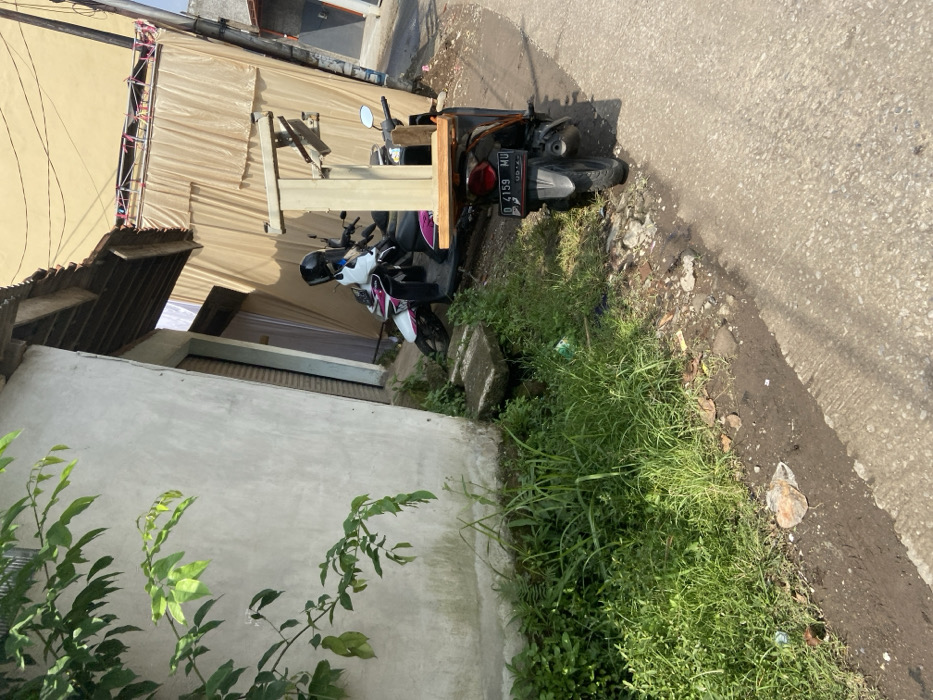 The image depicts a narrow street lined with concrete and paved with asphalt. A light-colored concrete building is visible on the left side of the street. There is a white and pink motorcycle with a license plate that reads "D 4159 MU" parked on the right side of the street. The motorcycle is next to a beige table with a wooden top and a metal frame, positioned perpendicular to the street. There is a second motorcycle, a white scooter with a black seat, partially obscured by the beige table. Lush, green foliage is visible on the left side of the street. The ground is covered with loose gravel and dirt. The image was taken in Margaasih, Indonesia, at coordinates -6.941 latitude and 107.552 longitude.