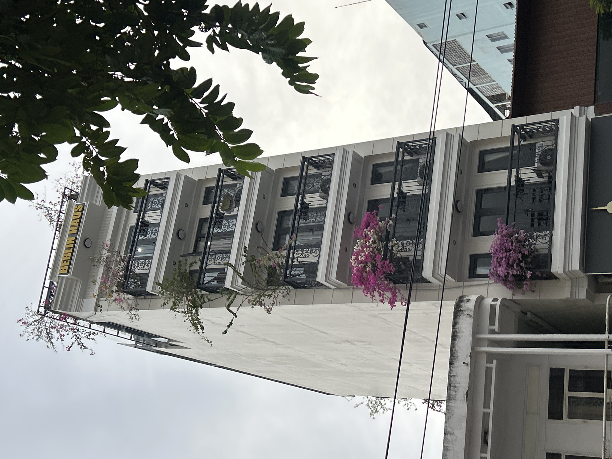 The image presents a multi-story building, likely a hotel or apartment complex, viewed from a low-angle perspective, partially framed by dark green tree leaves in the upper foreground. The sky is bright and uniformly overcast.
The building features a clean, white facade with several upper-floor balconies. These balconies are distinctively framed by white architectural molding and equipped with ornate black metal railings. Each balcony is adorned with vibrant clusters of flowers, predominantly pink and purple, with some white blooms, providing colorful accents. Some green foliage also trails from the balconies. Visible through the balcony railings and windows are air conditioning units.
On the upper left side of the building, a vertical sign displays the yellow text "BERLIN HAUS" on a dark background. Below the main building structure, a simpler, possibly older, white lower section or an adjacent building is partially visible.
In the upper right corner of the frame, a segment of another taller building with a lighter facade and a darker roof is seen. Multiple black power lines crisscross the upper part of the image, running diagonally across the sky and in front of the structures.
The scene depicts a static daytime urban environment under an overcast sky. The overall setting is consistent with an urban area like Nha Trang Ward, Vietnam, as provided by the location context.