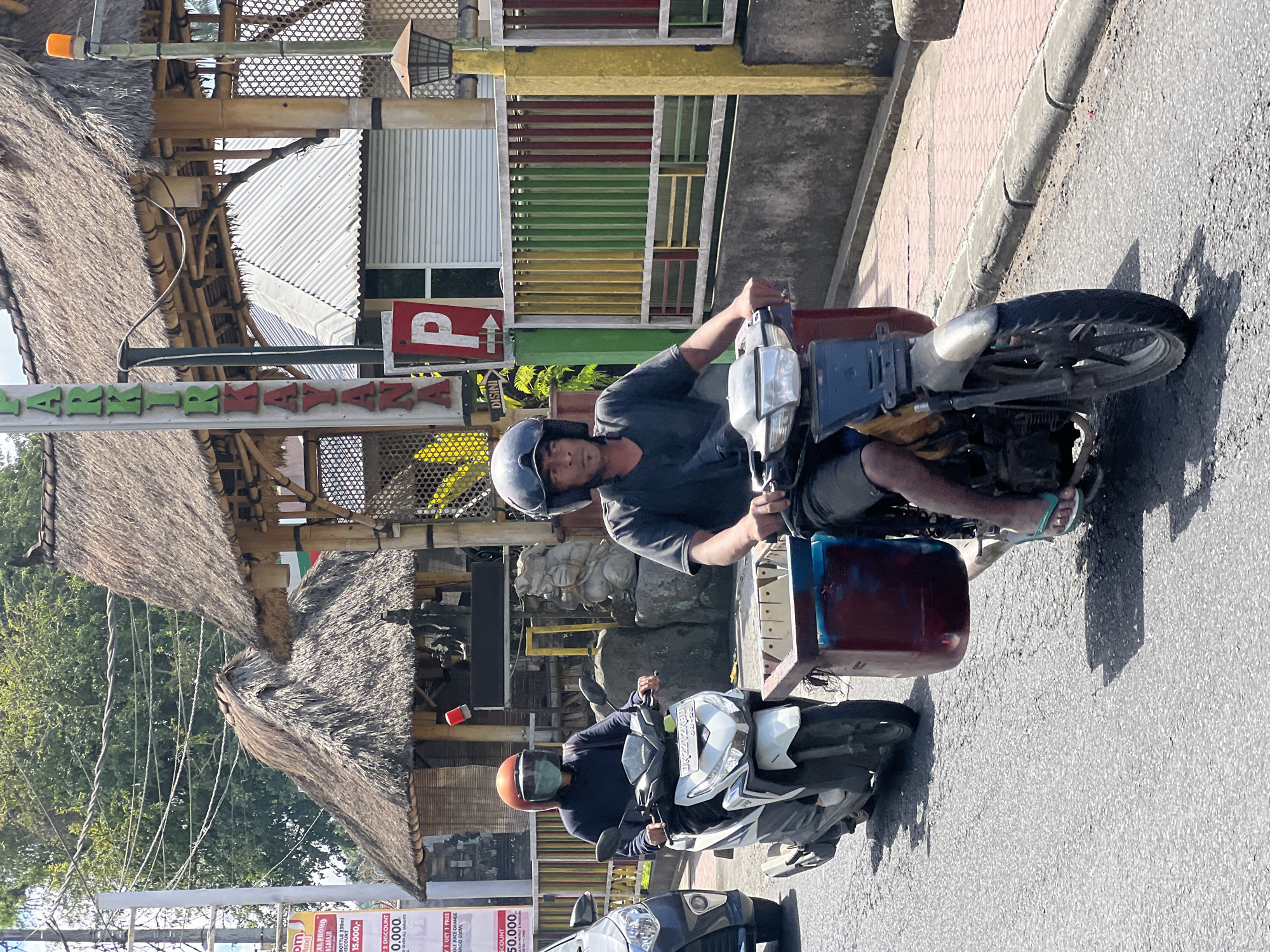 In Ubud District, Indonesia, two individuals are riding motorcycles on a paved road. The rider in the foreground, wearing a dark t-shirt and helmet, is operating a motorcycle with a large red and blue container attached to the front. The rider behind them, wearing a dark hoodie and an orange helmet with a green face shield, is on a white scooter. The background features traditional Balinese architecture with thatched roofs, a colorful fence, and a sign that reads "PARKIR KAYAANA". The scene is lit by bright daylight, casting shadows on the road.