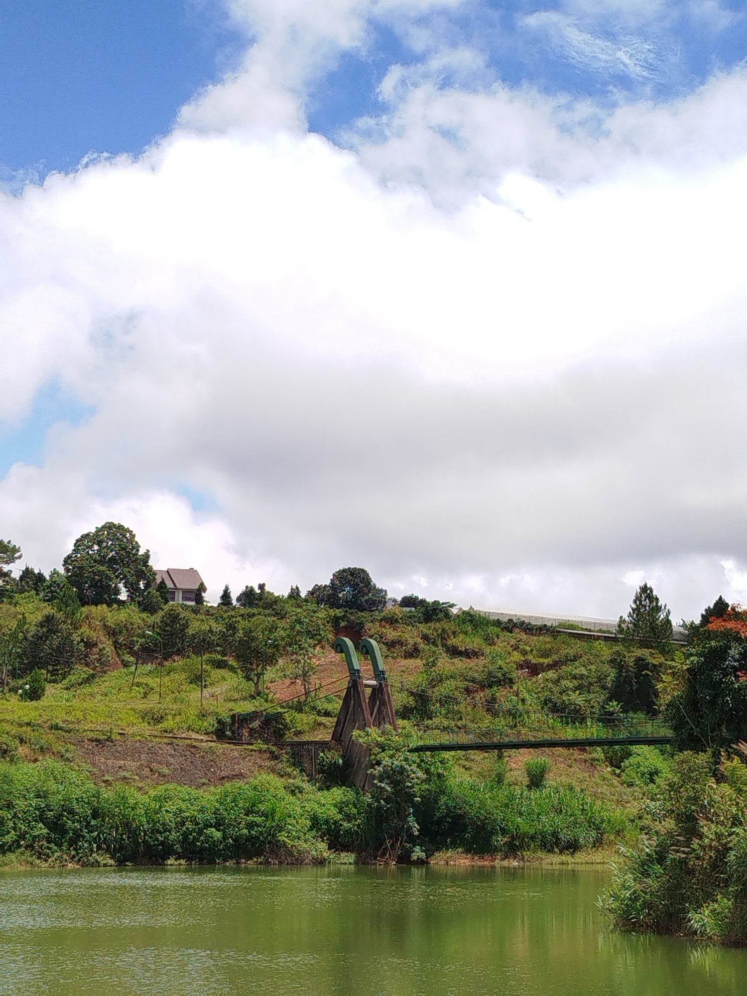 The image depicts a serene natural landscape with a body of water in the foreground and a verdant hillside in the background. The water, a tranquil green, reflects the soft light of the sky, which is a mixture of bright blue and fluffy white clouds. The hillside is covered in lush vegetation, with a variety of trees and shrubs creating a textured, layered effect.

Near the crest of the hill, a charming house with a brown roof is nestled amongst the trees, suggesting a dwelling in a rural or semi-rural setting. Further along the hillside, a notable feature is a set of large, green pipes that appear to be part of an industrial or water management system, possibly for irrigation or drainage. Adjacent to these pipes, a narrow, green-painted bridge spans a small gap, hinting at a pathway or access route. The overall impression is one of a picturesque and somewhat remote location, possibly a park, a nature reserve, or a private estate.

The location is identified as being in the city of Lạc Dương, Vietnam. Lạc Dương is known for its natural beauty, often characterized by rolling hills, pine forests, and cool climates, which aligns with the visual elements present in the photograph. The scene likely captures a specific point of interest within this area, perhaps a reservoir or a landscaped viewpoint that highlights the region's natural appeal alongside human-made structures.